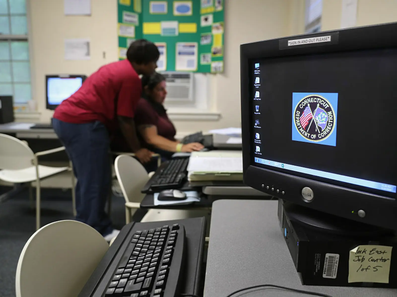 A computer monitor is seen in the foreground as a woman helps another at a computer in the background.