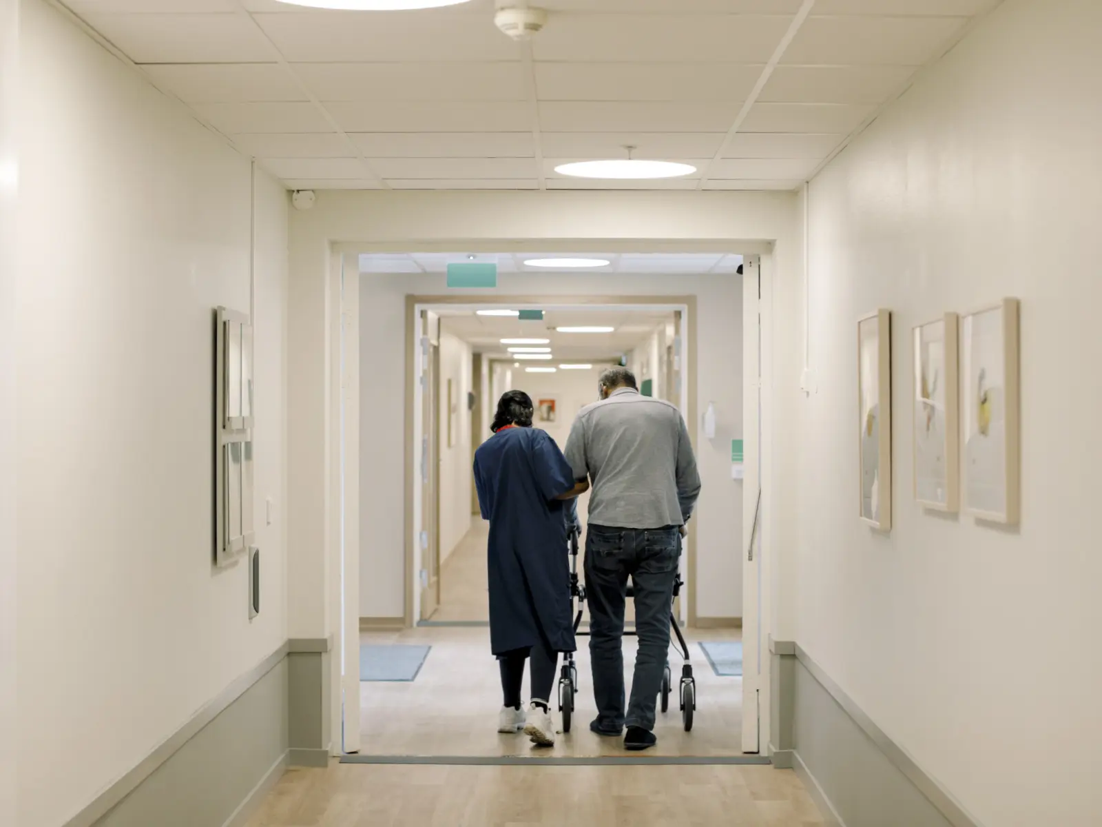 A caregiver assists an elderly person with a walker down a light-colored corridor, framed by soft, ambient lighting and artwork.