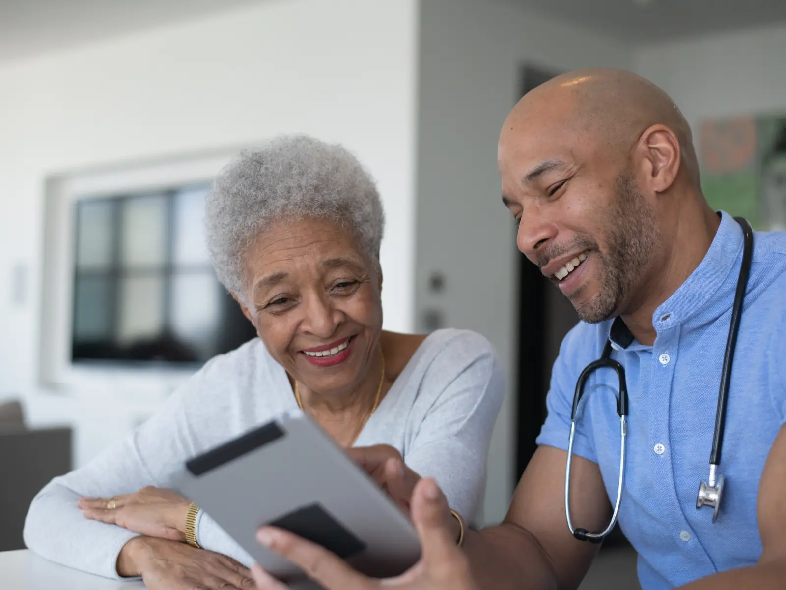 A smiling, older woman and a male doctor share a joyful moment while looking at a tablet in a modern, bright environment.
