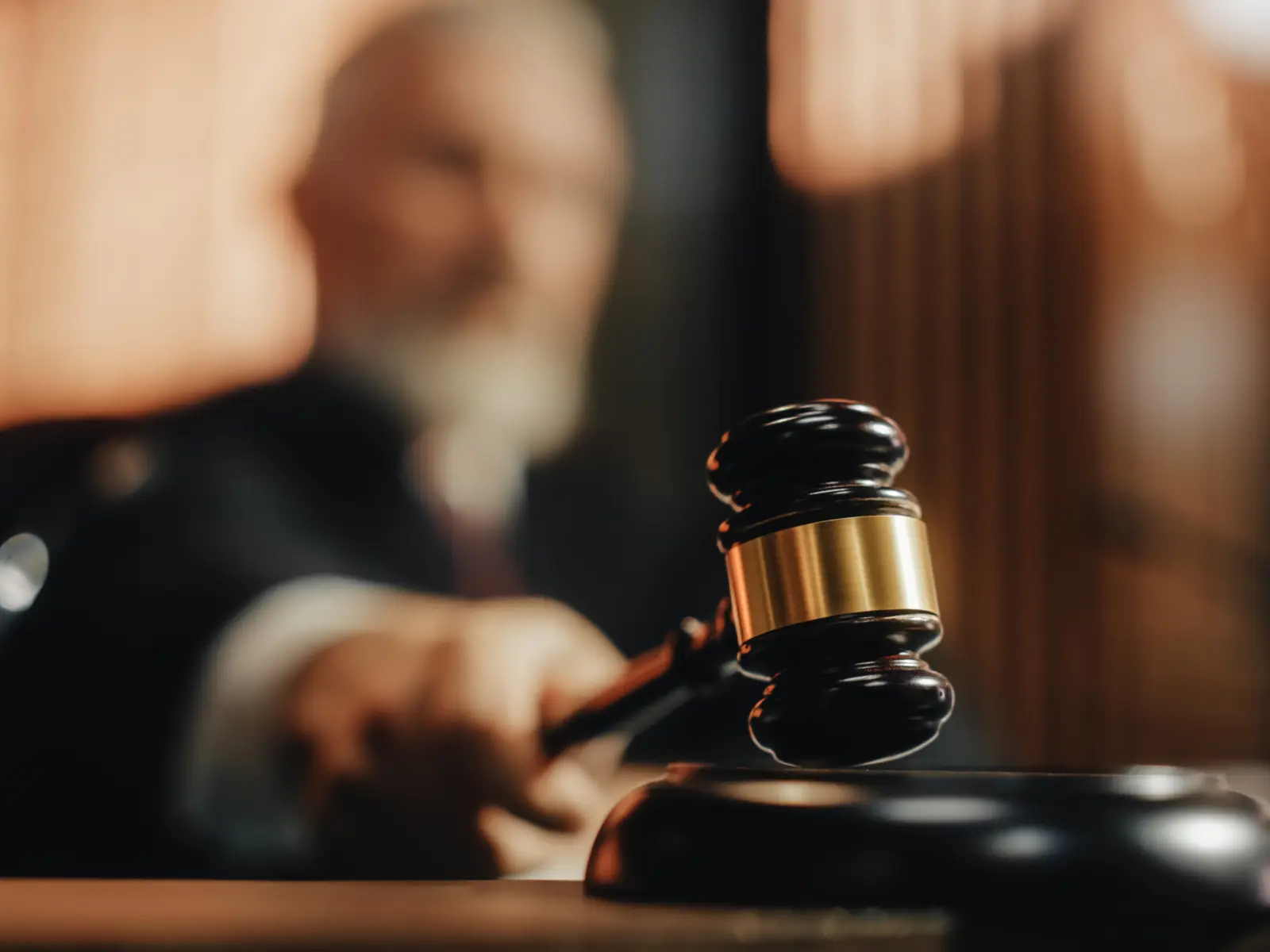 A gavel is poised for a strike on its base, with a blurred judge in the background, evoking a courtroom setting.