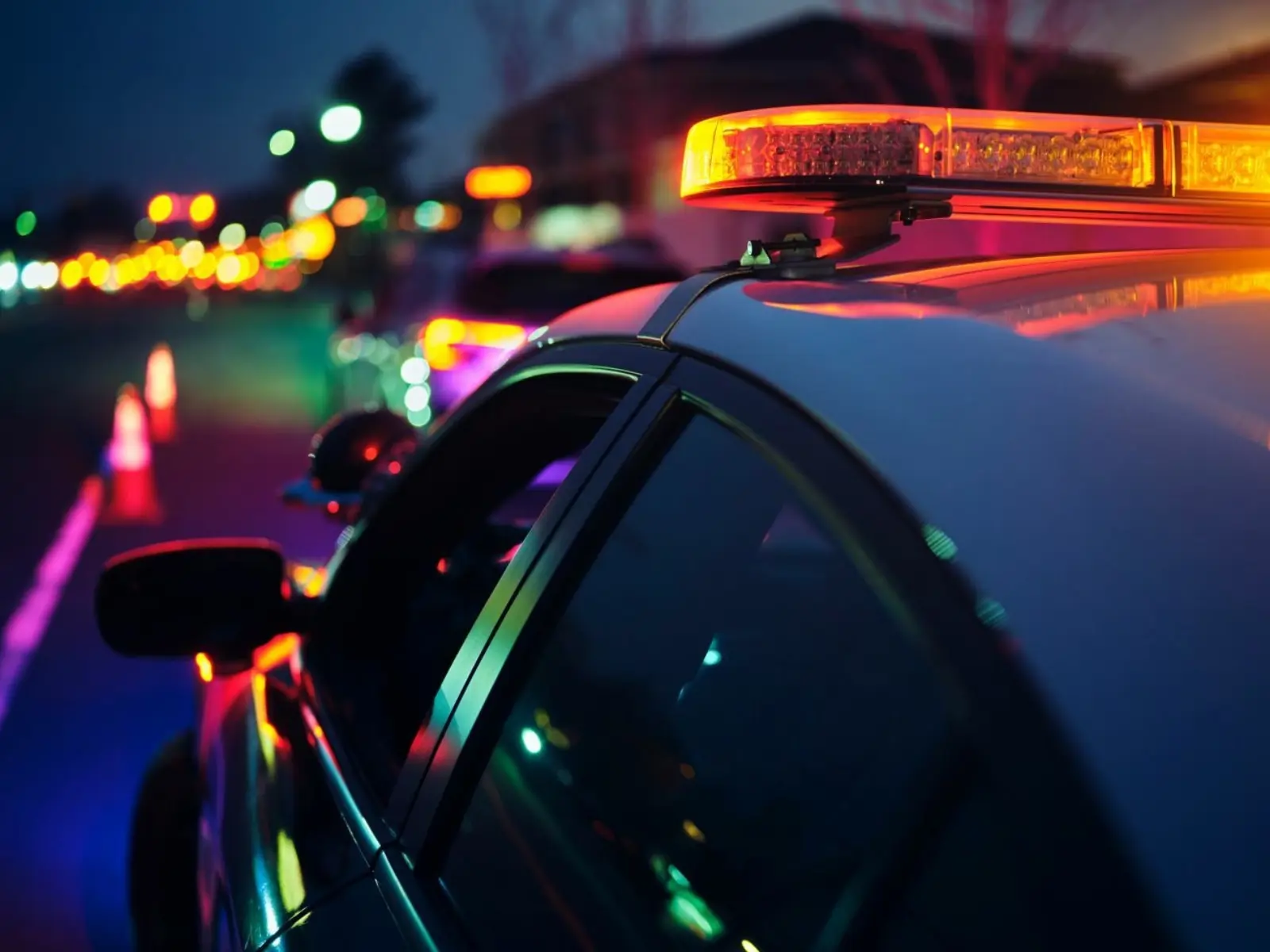A police car with illuminated sirens is shown at night, reflecting colorful lights on nearby vehicles and traffic cones.