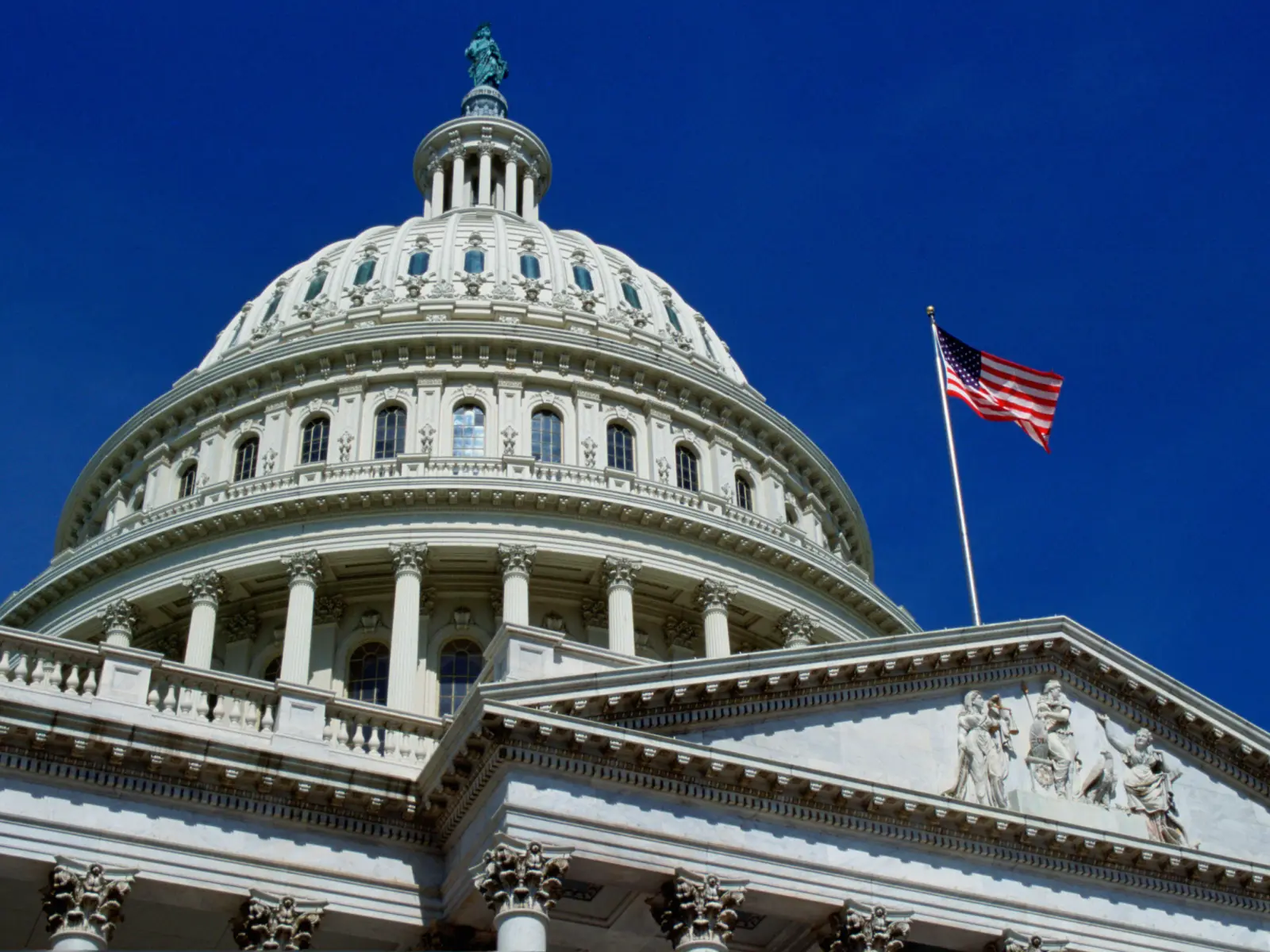 The U.S. Capitol dome rises against a clear blue sky, topped by a statue, with an American flag waving on a columned building.
