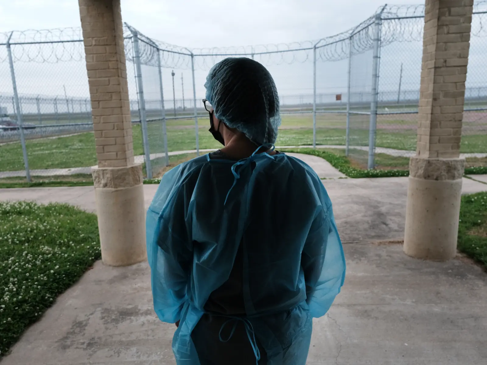 A person wearing protective blue medical gear stands at an entrance, gazing out toward a fenced area with grass and barbed wire.