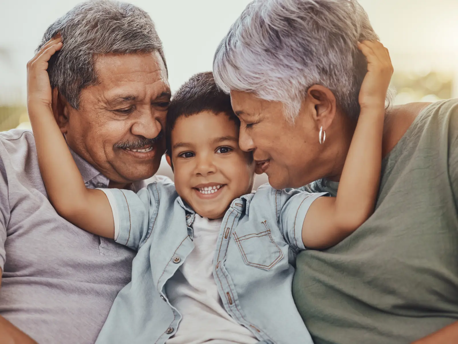 A smiling boy sits between his joyful grandparents, sharing a warm embrace, radiating love and connection in a sunlit setting.