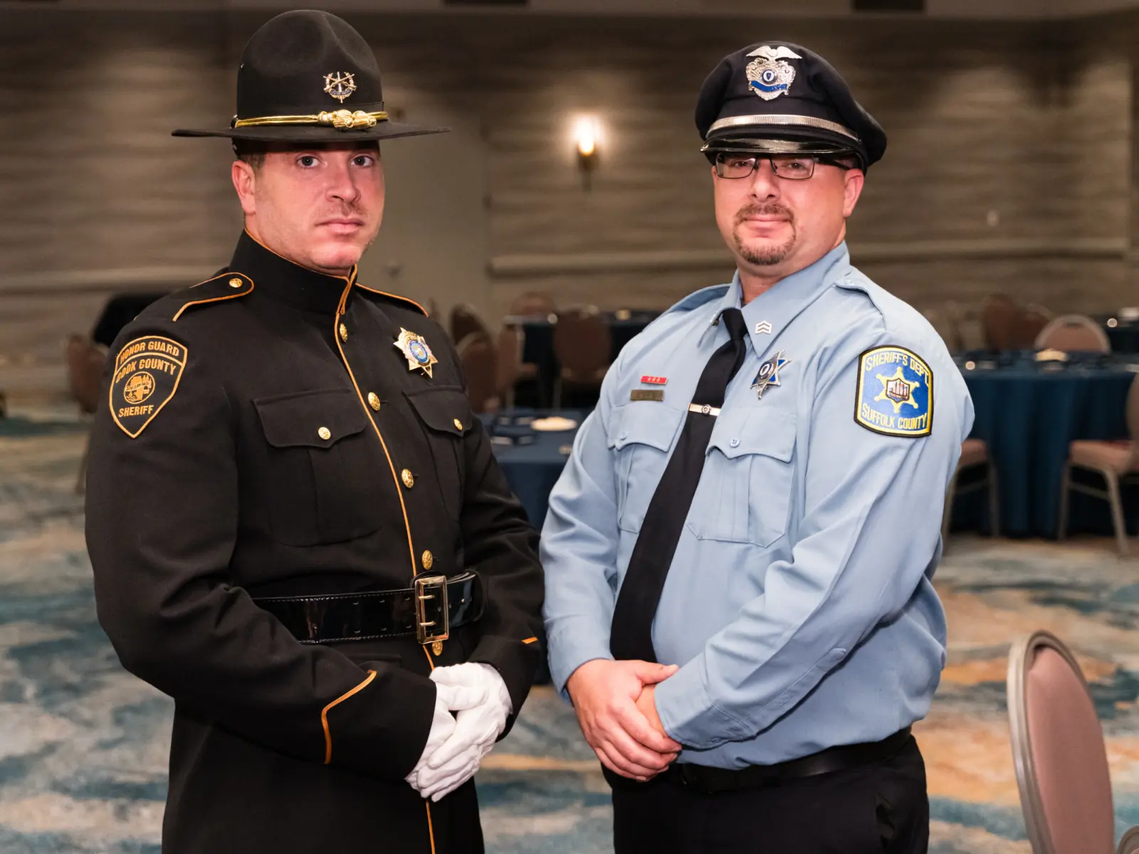 Two law enforcement officers stand together in a banquet hall, one in a formal dark uniform with a hat, the other in a light blue uniform.