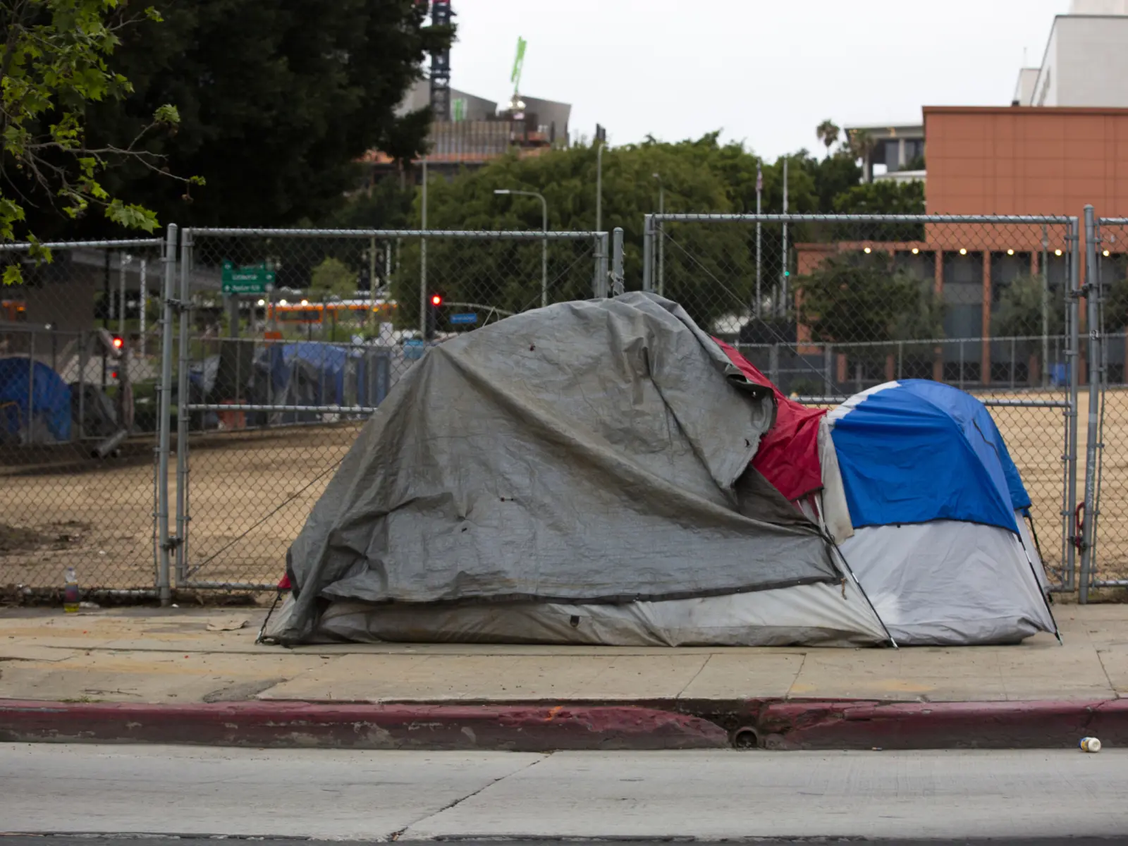 A weathered tent, covered with a gray tarp and touches of red and blue, sits along a sidewalk near a fenced area.