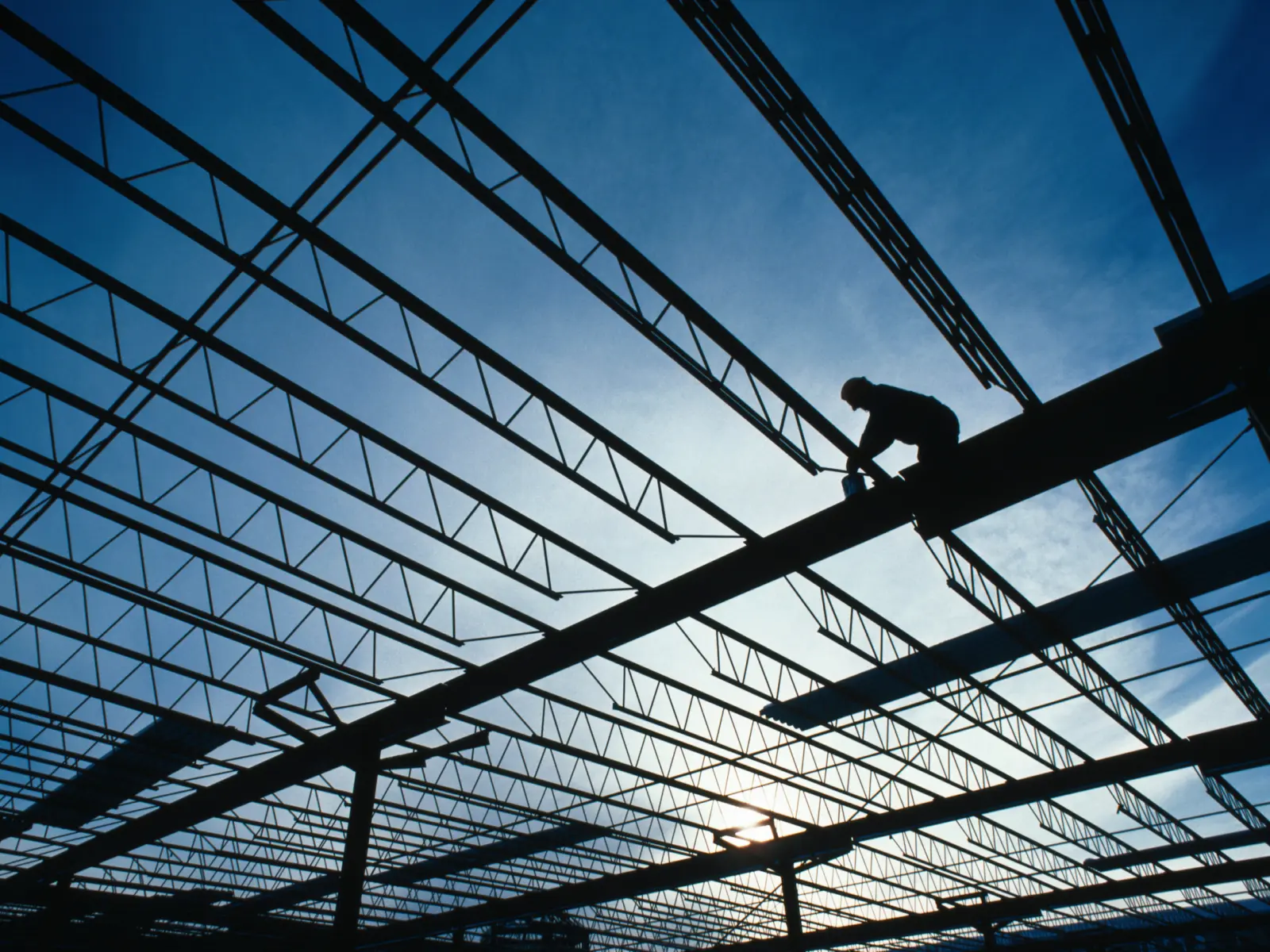 A silhouetted worker balances on a steel beam, gripping a tool, against a blue sky and intricate framework of beams above.