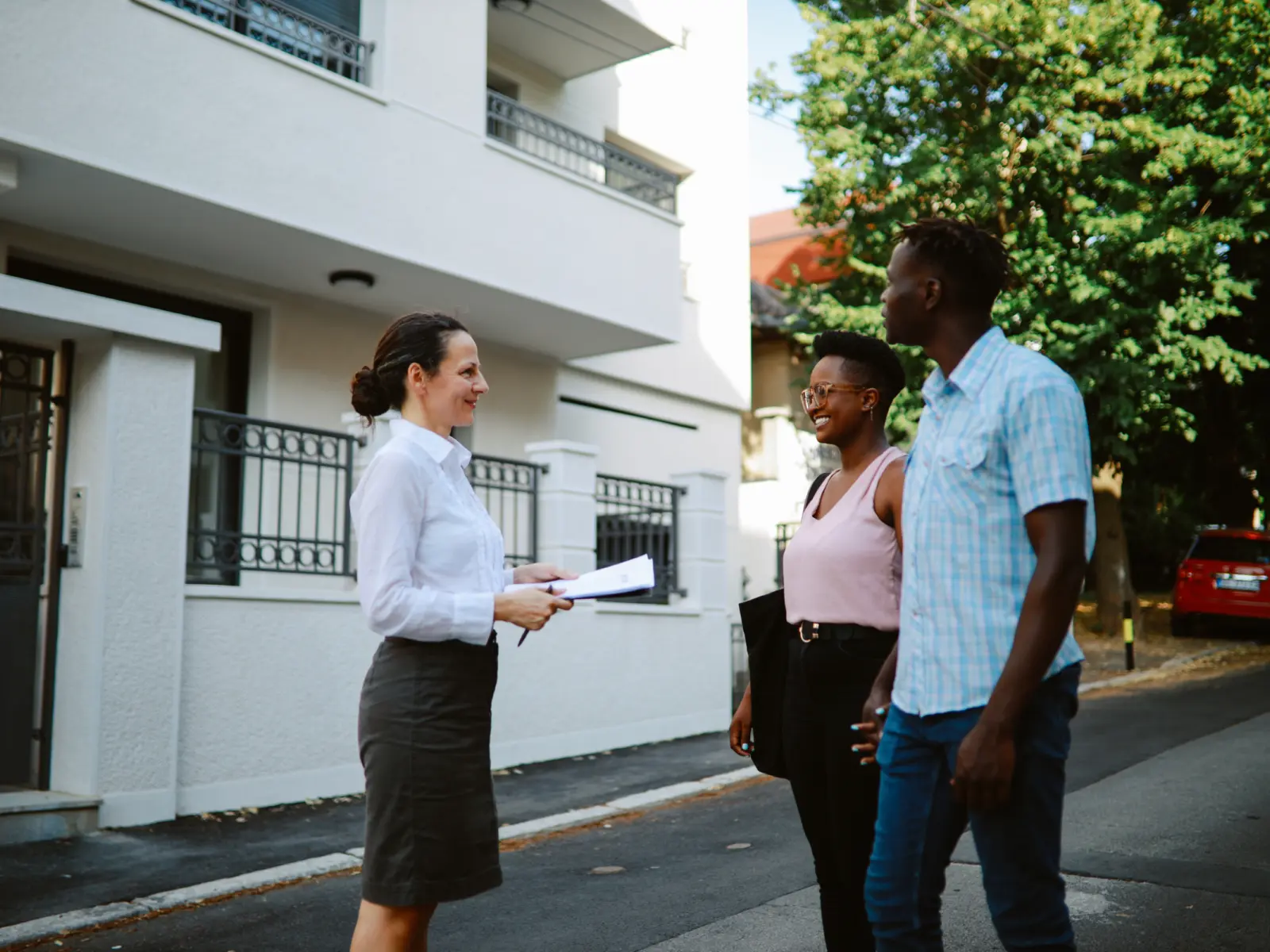 A woman in a white shirt and skirt holds papers while smiling at two people on a street, surrounded by greenery and buildings.