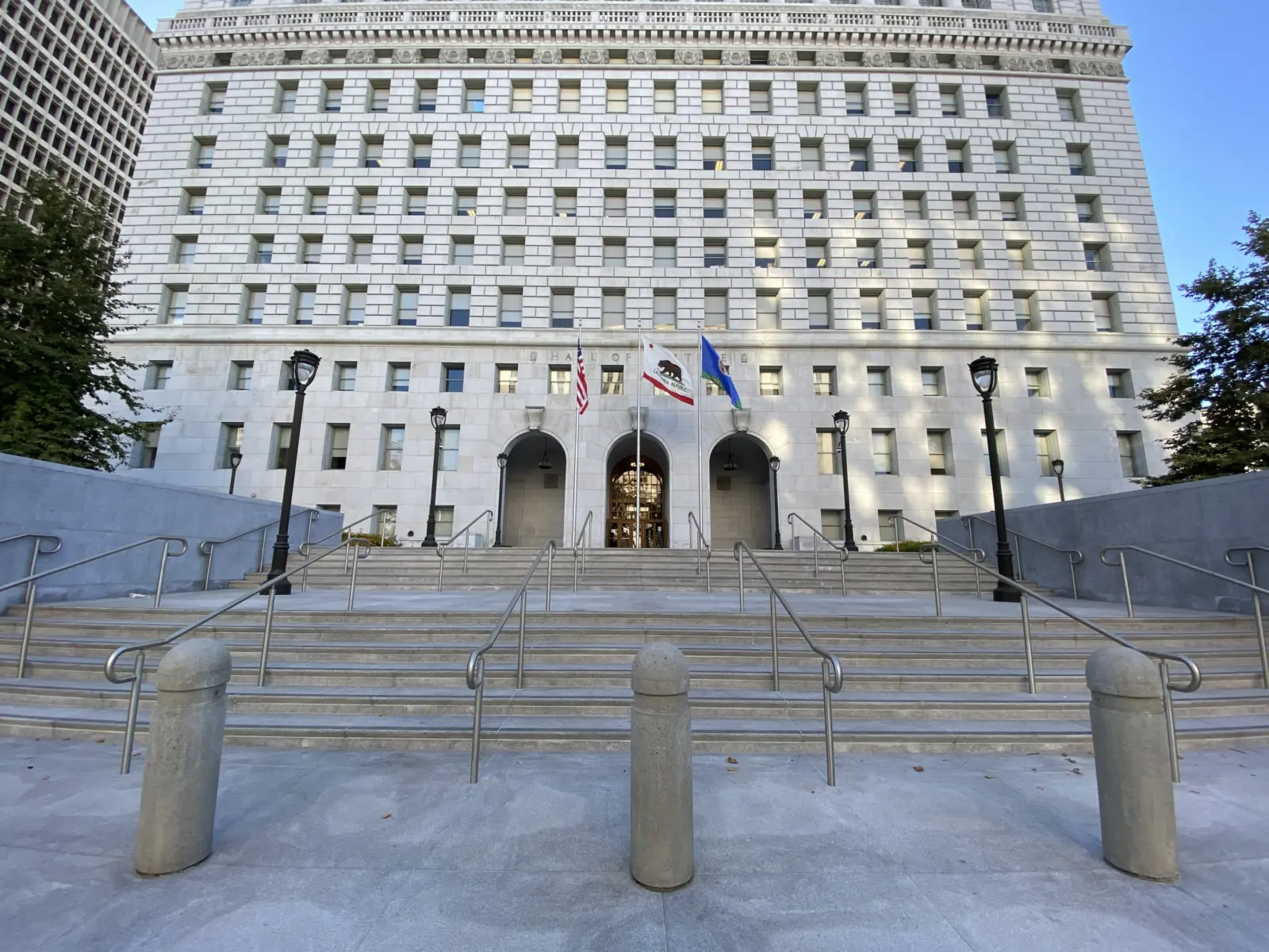 A grand stone building with numerous windows features a wide staircase, flanked by lampposts and flags at the entrance.