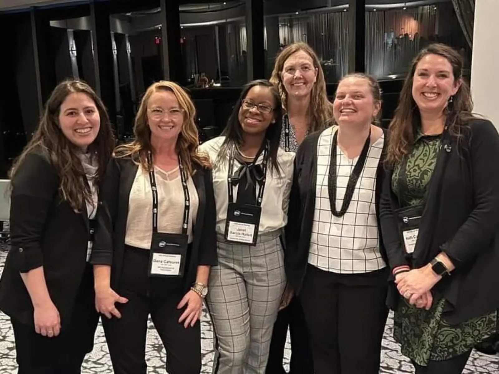 Six women pose together in a conference setting, wearing badges, smiling, and dressed in professional attire against a stylish backdrop.