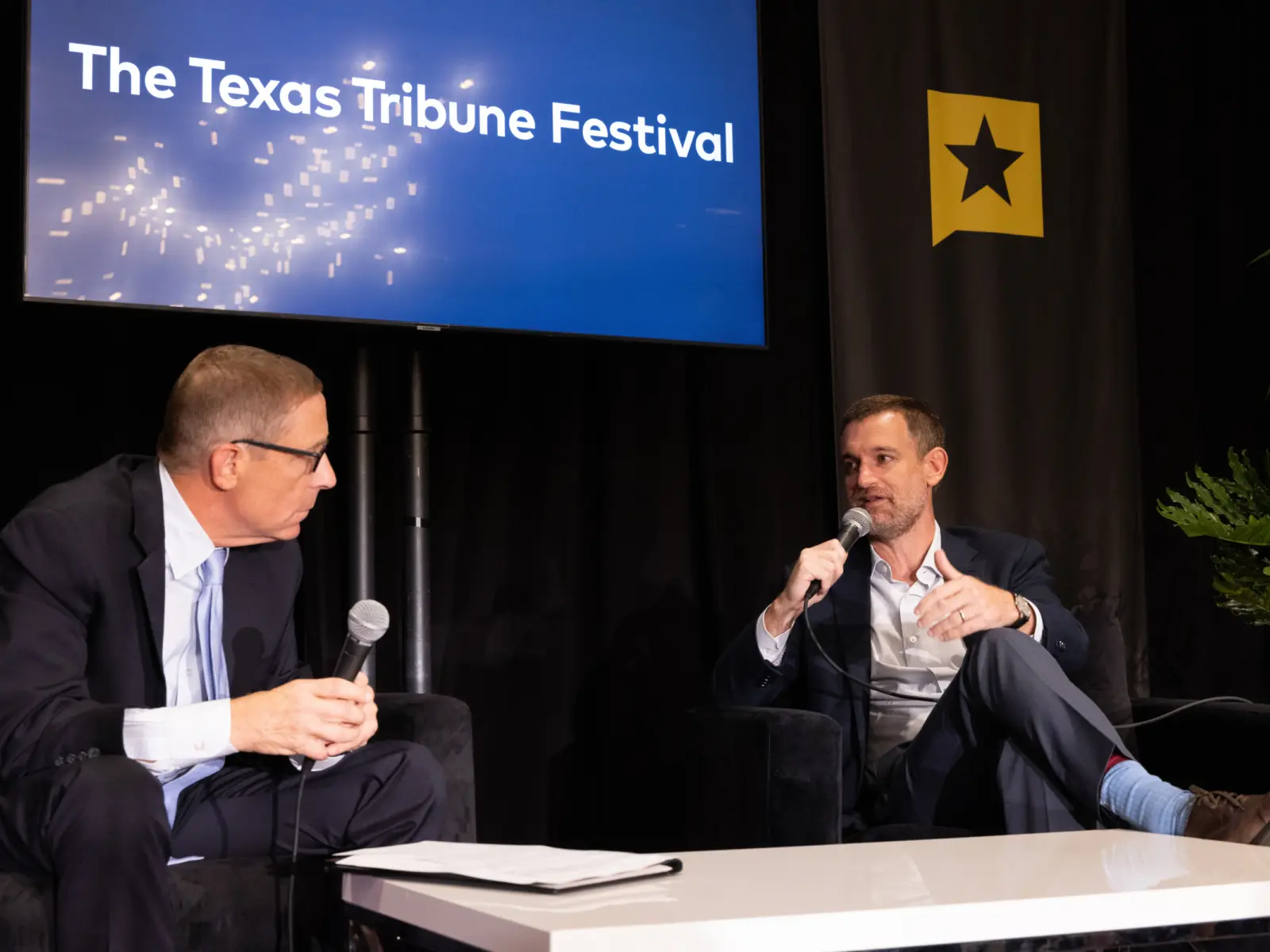 Two men, one speaking into a microphone, engage in conversation on stage at the Texas Tribune Festival, with a spotlighted backdrop.