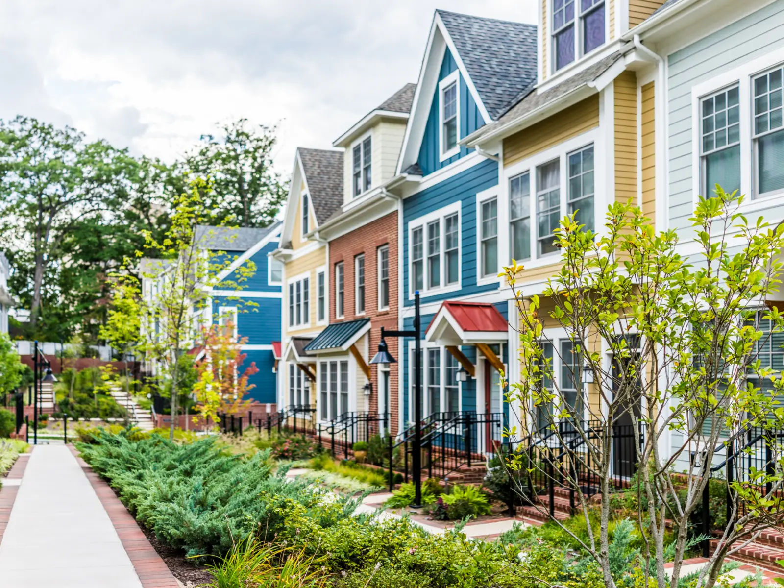 Colorful townhouses line a well-maintained path, surrounded by greenery and blooming plants, under a partly cloudy sky.