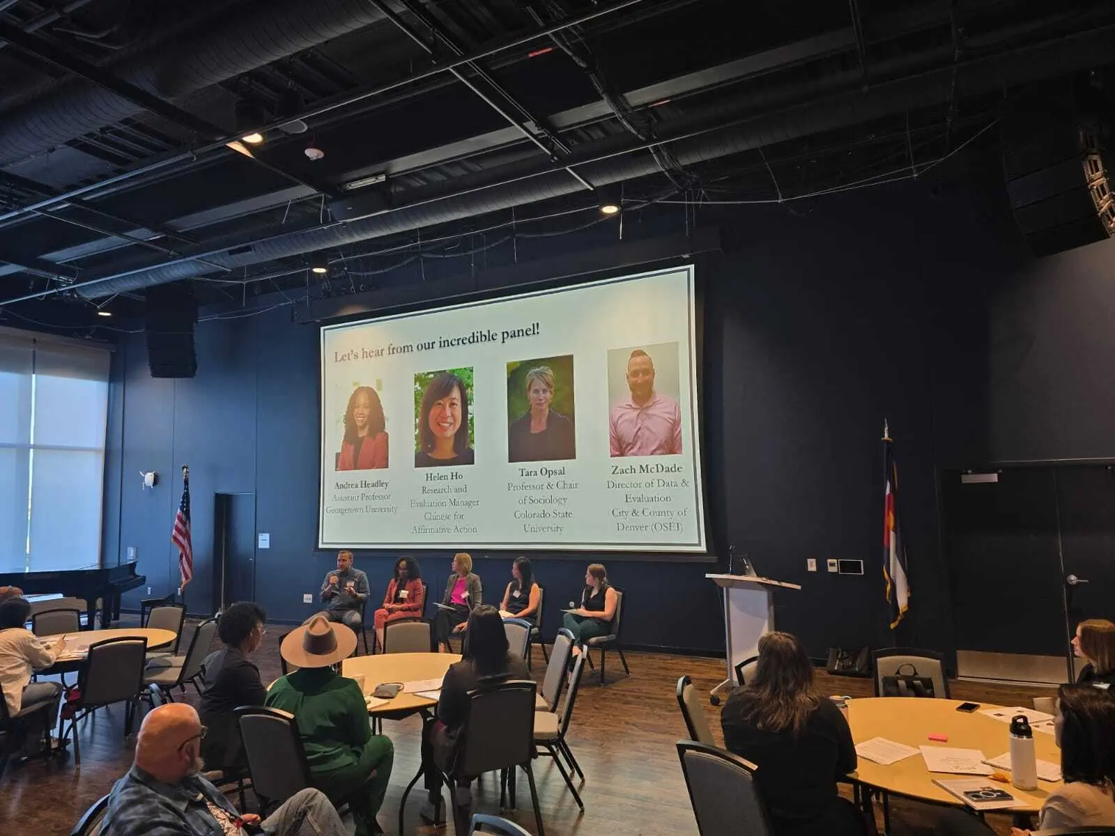 A panel discussion featuring four speakers is taking place in a spacious room with round tables and a large screen displaying their names.