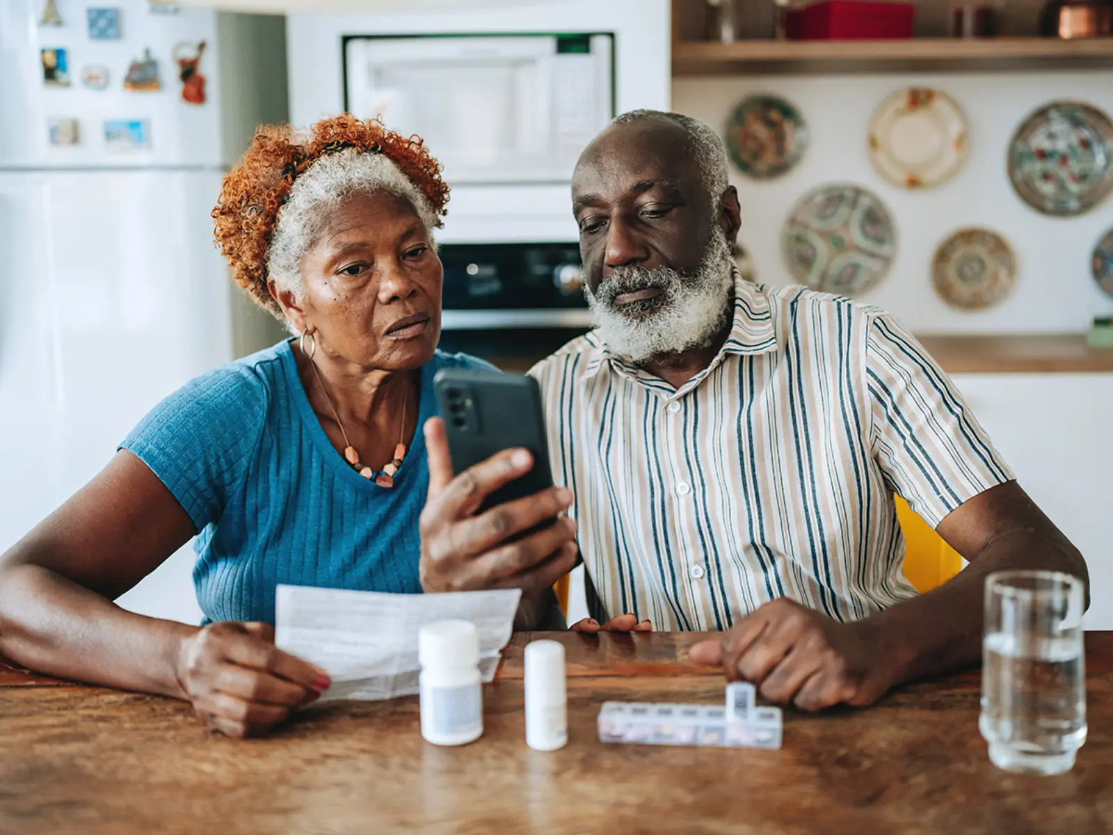 An older couple sits at a wooden table, reviewing a medication list on a phone, surrounded by prescription bottles and a glass of water.