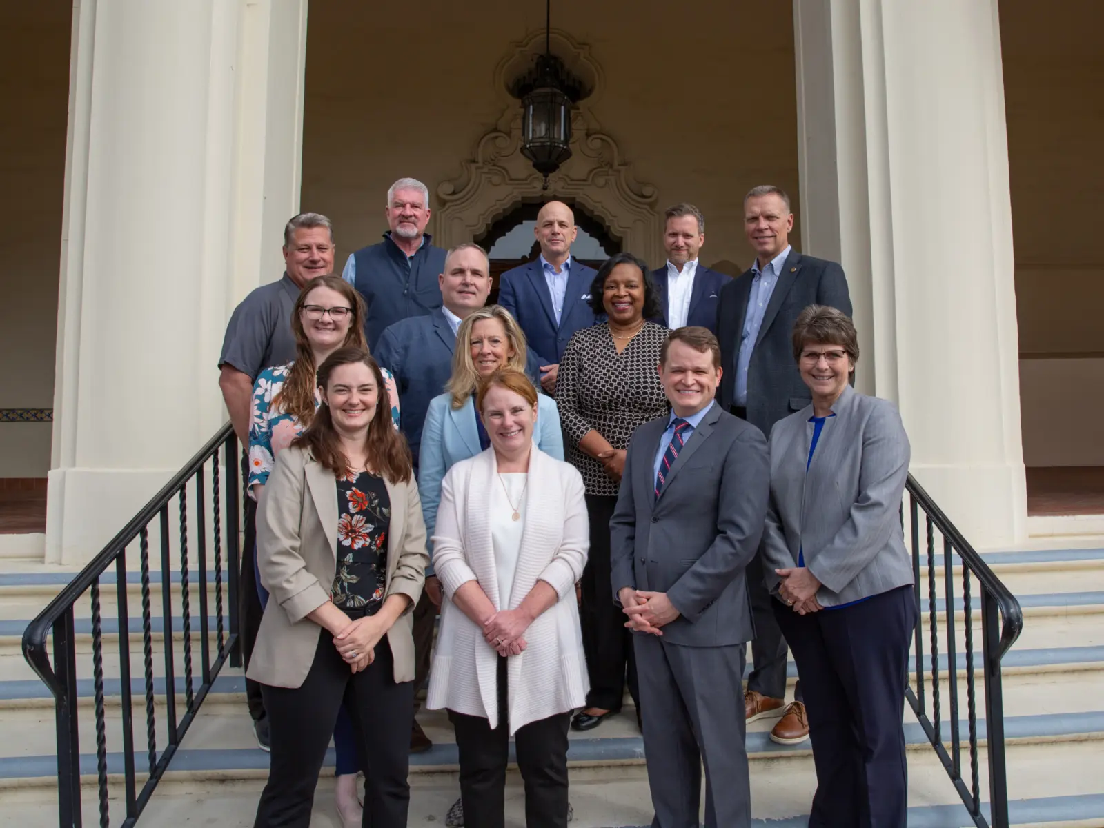 A diverse group of 15 people stands on stone steps, posing outdoors in front of a decorative building with a large lantern above.