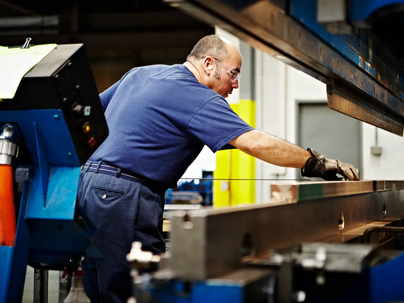 A worker in safety glasses operates a large metal bending machine, focusing intently on precise metal forming in a manufacturing setting.
