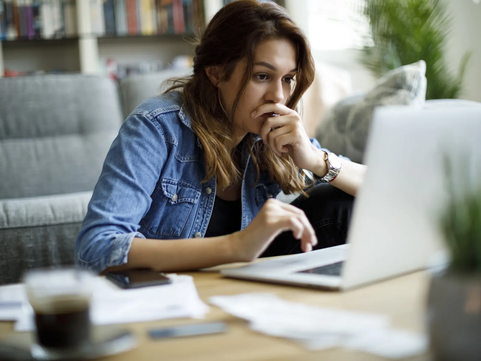 A young woman with wavy hair sits at a table, focused on her laptop, hand on chin, surrounded by papers and a coffee cup.