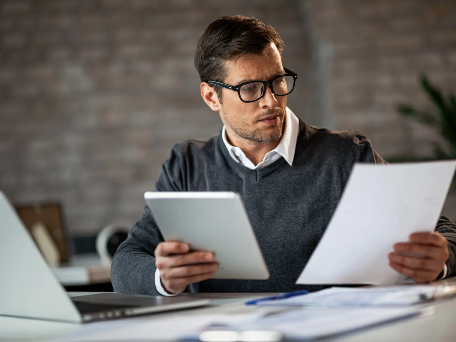 A man in glasses examines a document while holding a tablet, seated at a desk with a laptop and papers in an office.