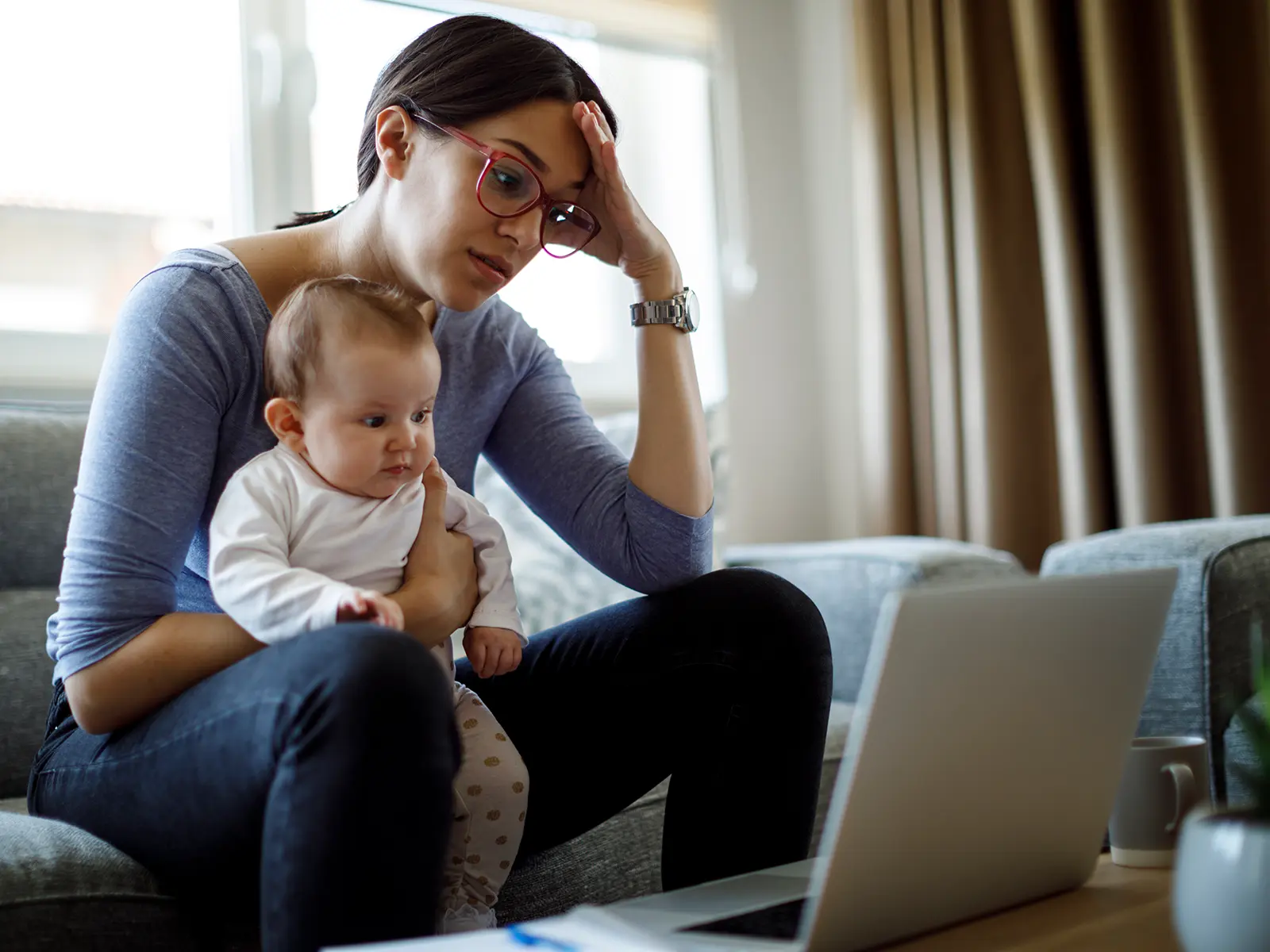 A woman sits on a couch, holding an infant, looking stressed while working on a laptop.