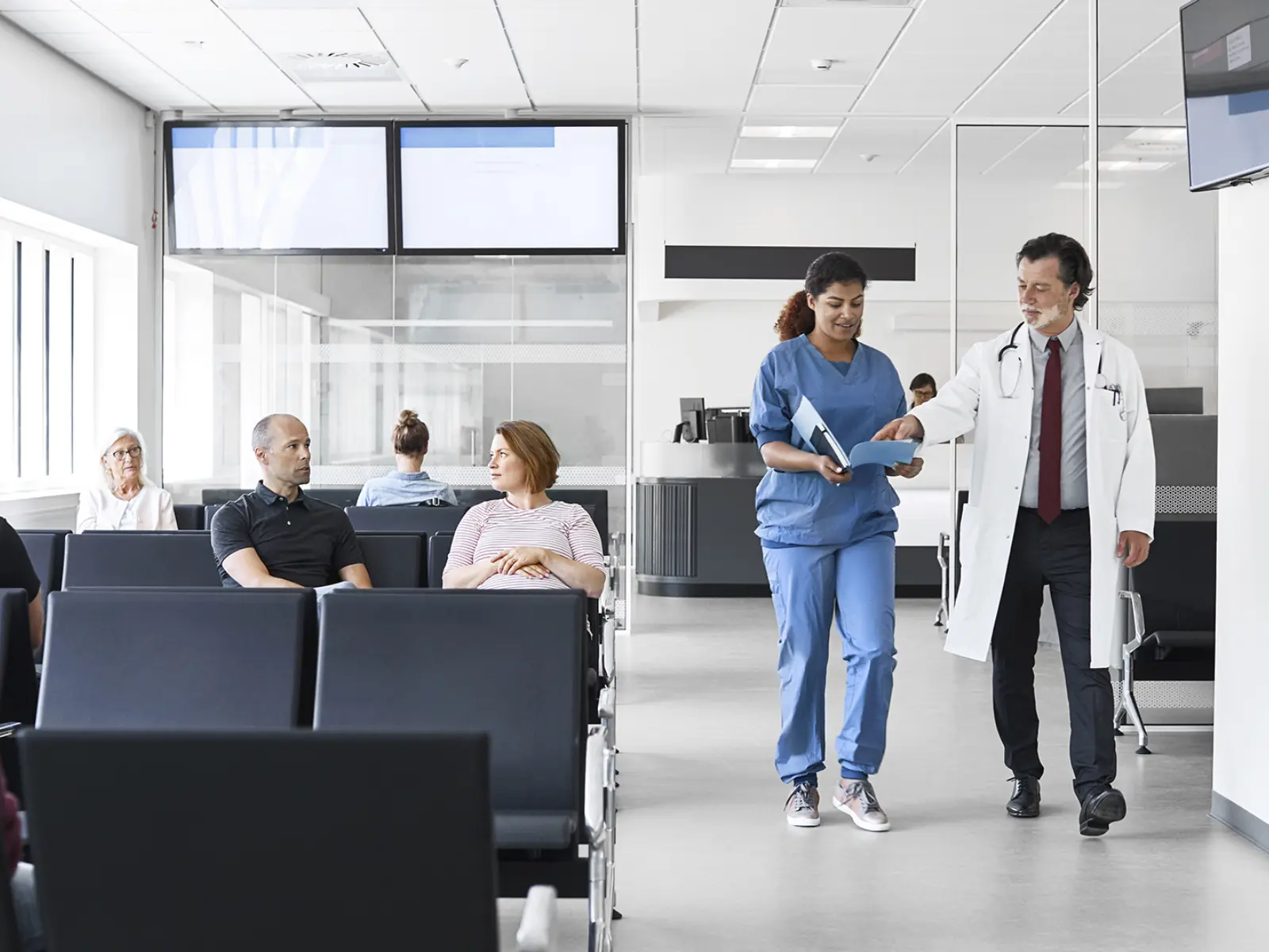A nurse in scrubs walks with a doctor in a white coat, discussing a patient file in a modern hospital waiting area.