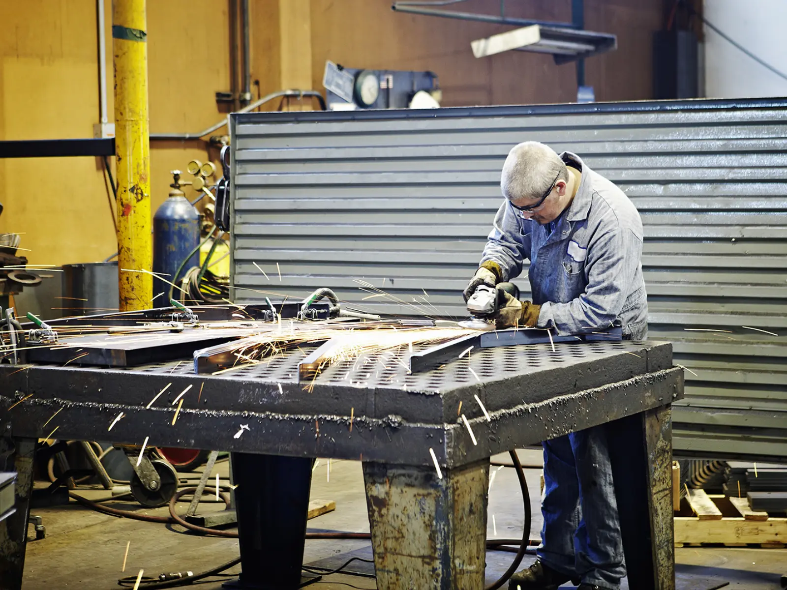 A worker in protective gear uses a grinder on a metal workpiece, surrounded by tools and sparking metal shavings in a workshop.