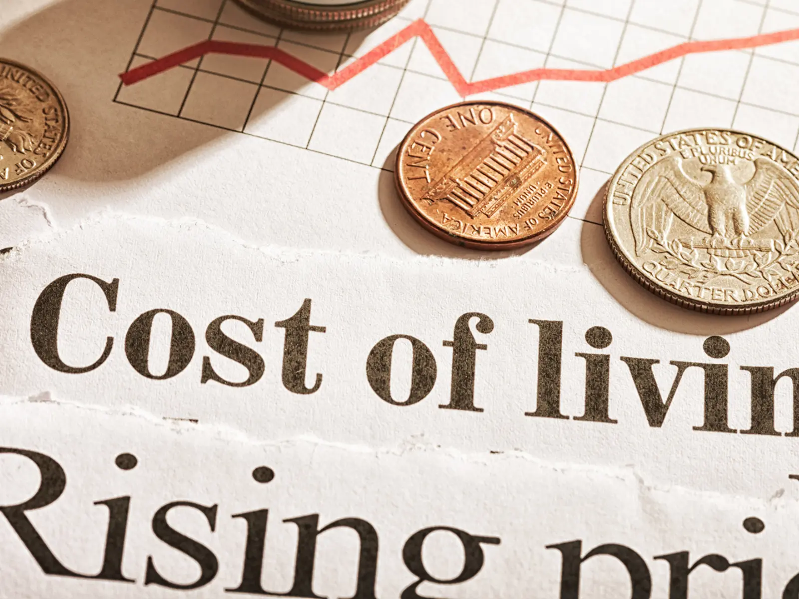 Coins are placed on torn newspaper headlines discussing the rising cost of living and prices, alongside a red line graph.
