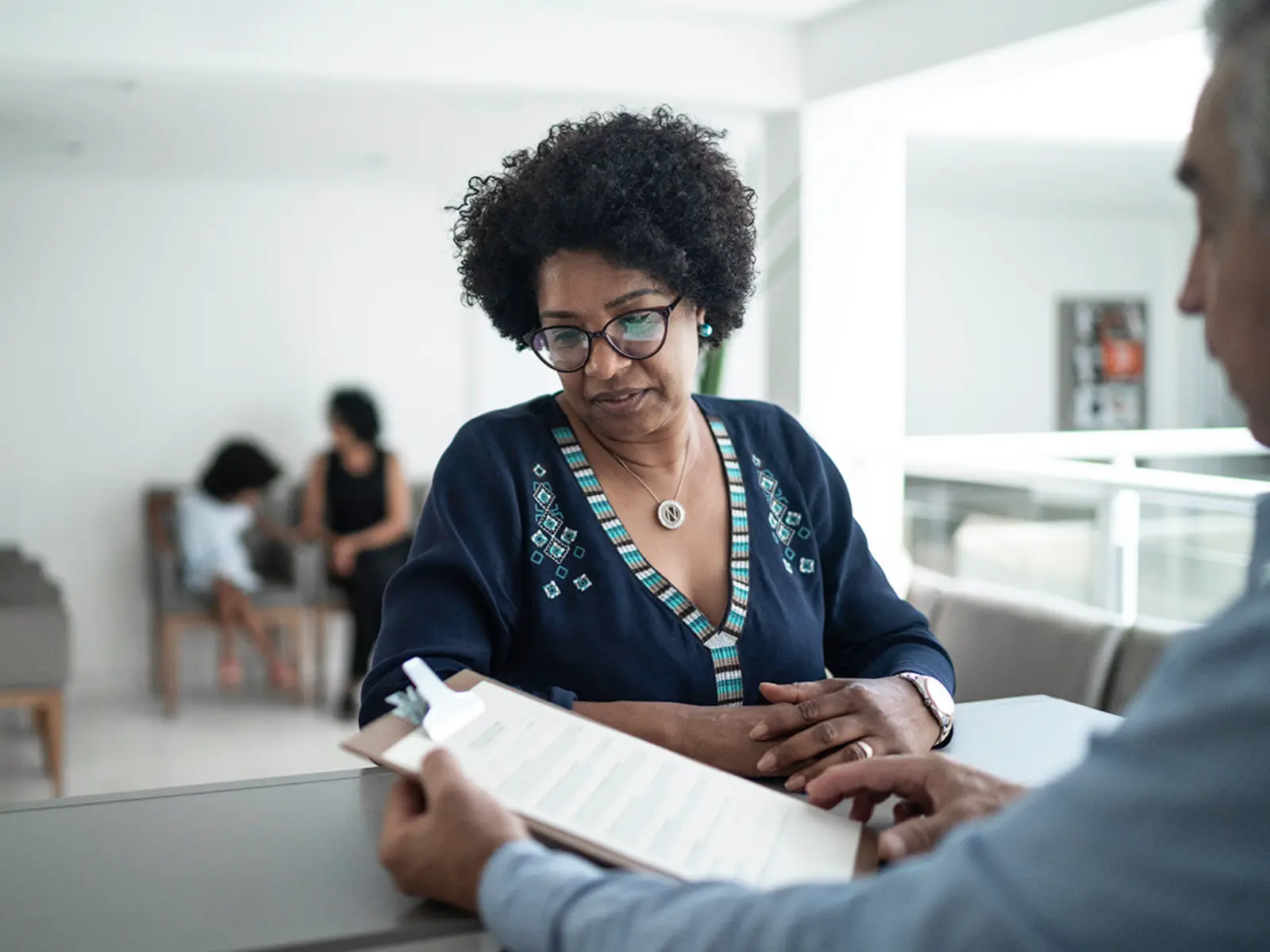 A woman with curly hair and glasses looks thoughtfully at a document presented to her, while a man gestures during a conversation.