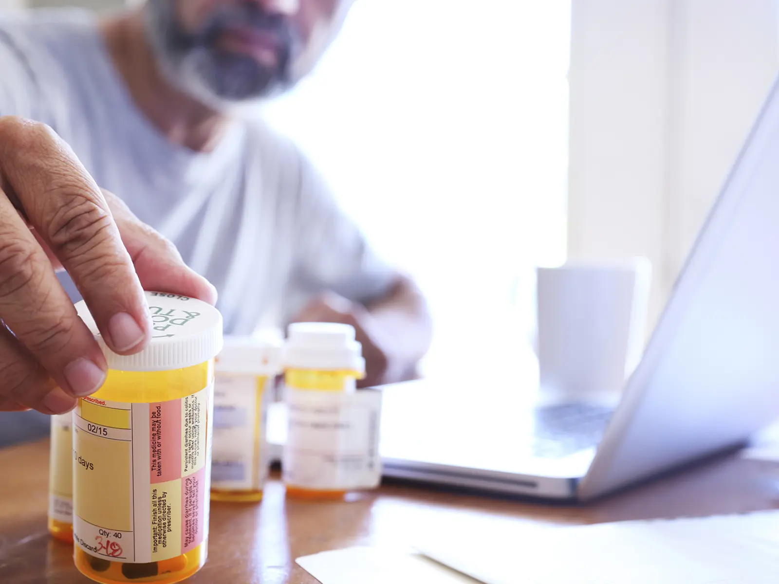 A person reaches for a prescription bottle on a wooden table, with other bottles and a laptop visible in the background.