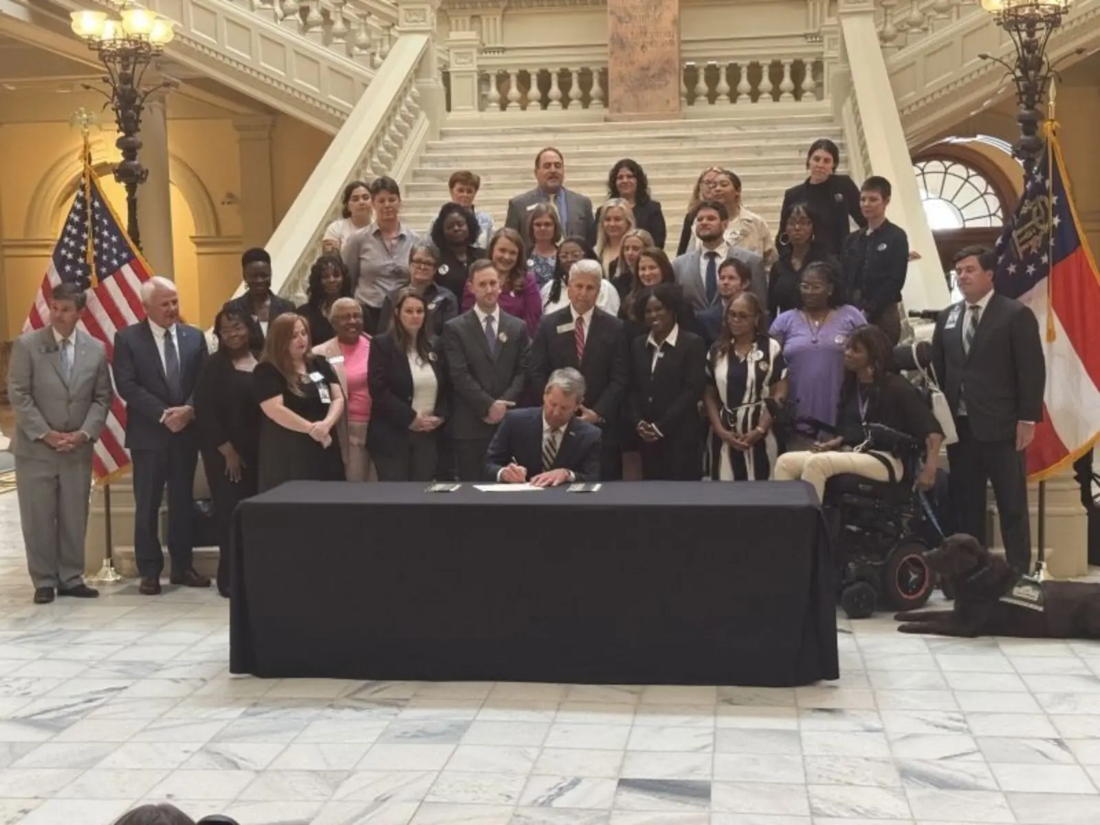 A group of diverse individuals gathers around a table as a man signs a document, with flags and an ornate staircase in the background.