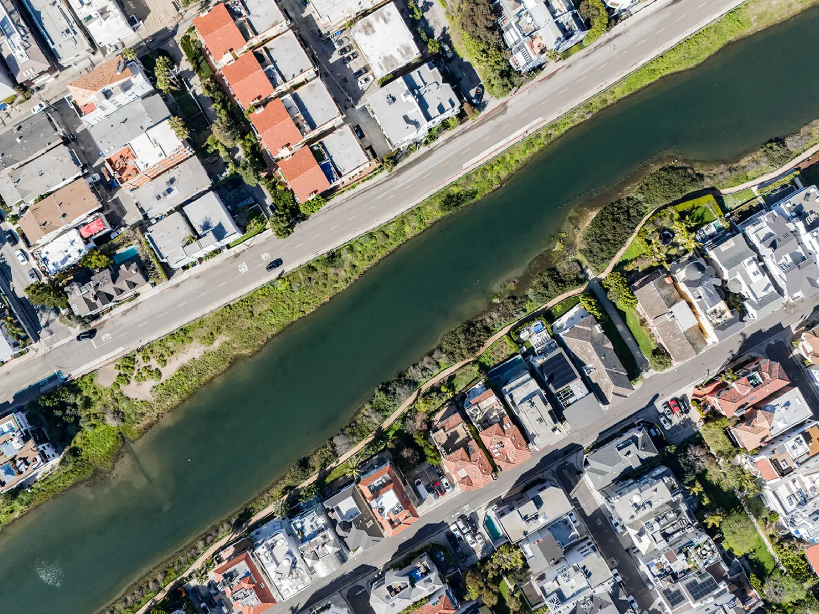 Aerial view of a residential area with colorful houses lining a green canal, bordered by a road and vegetation on both sides.
