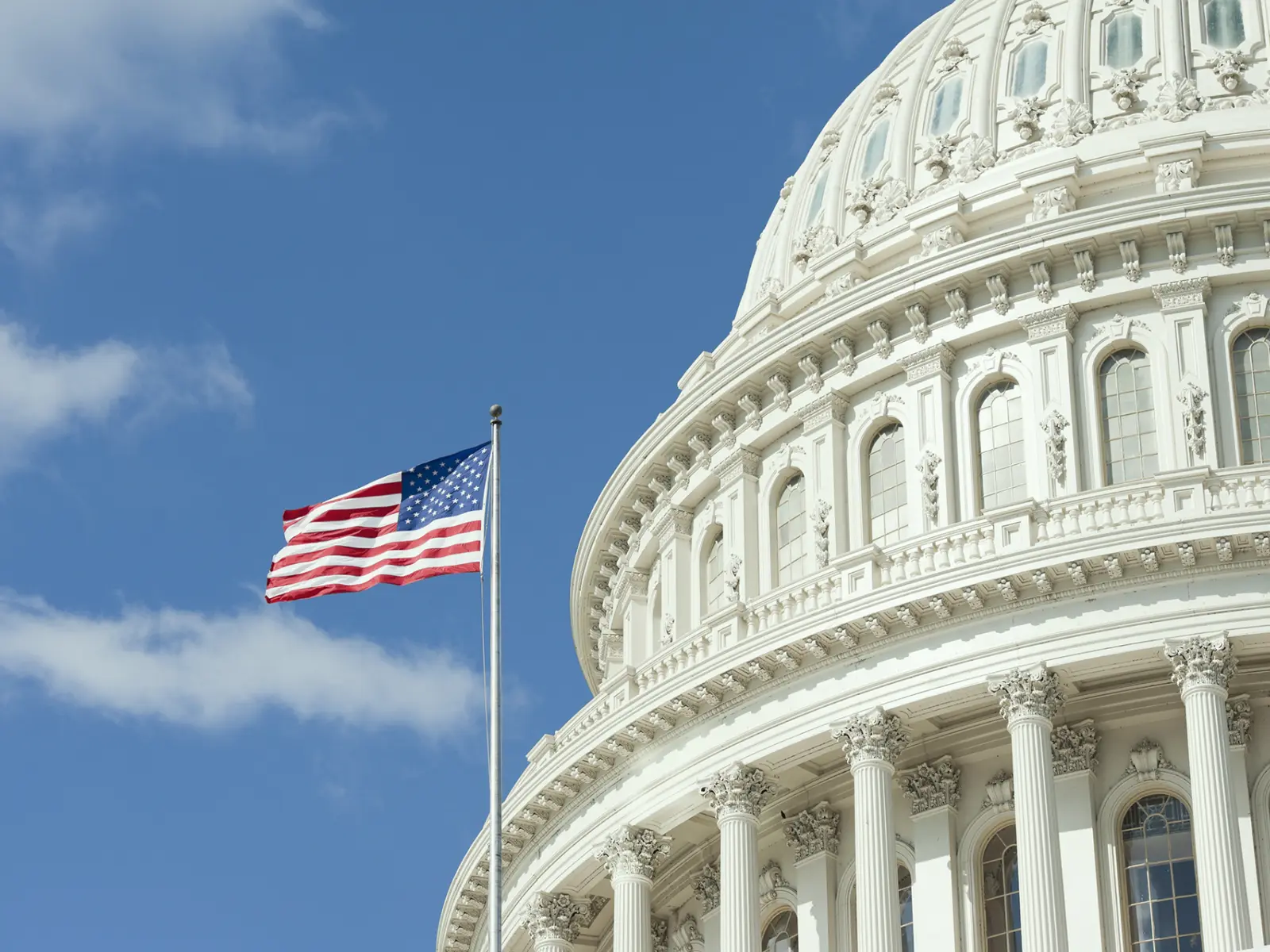 The U.S. Capitol dome partially captures the bright blue sky, with the American flag waving in the foreground.