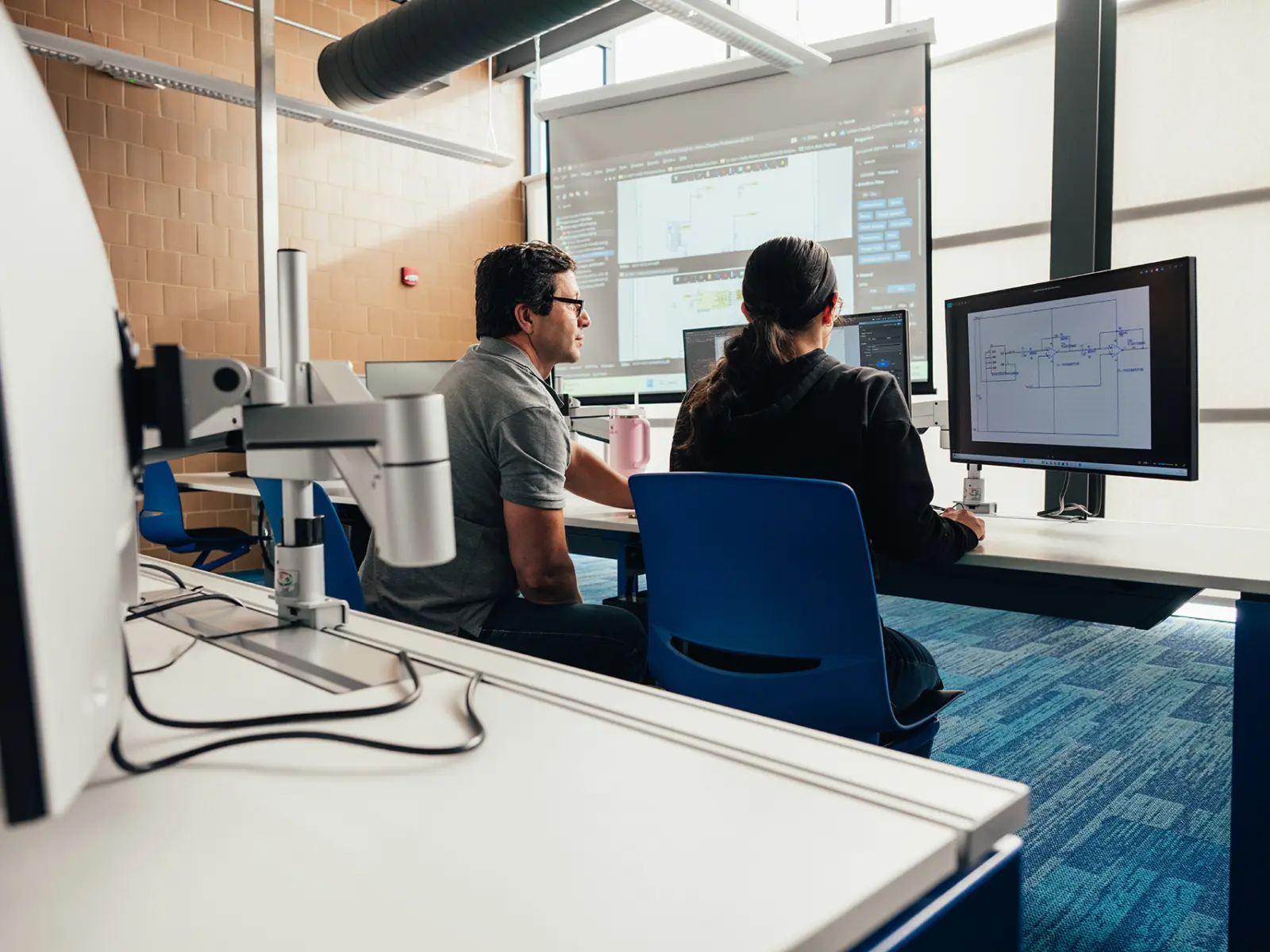 Two individuals focus on their computer screens in a modern classroom, with diagrams displayed on their monitors and a projector in use.