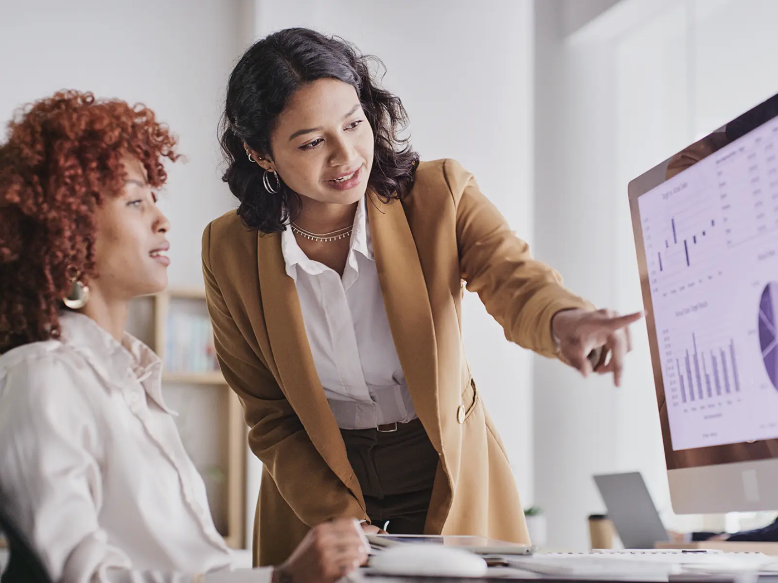 A businesswoman in a brown blazer points at data graphs on a computer screen while discussing insights with a colleague.