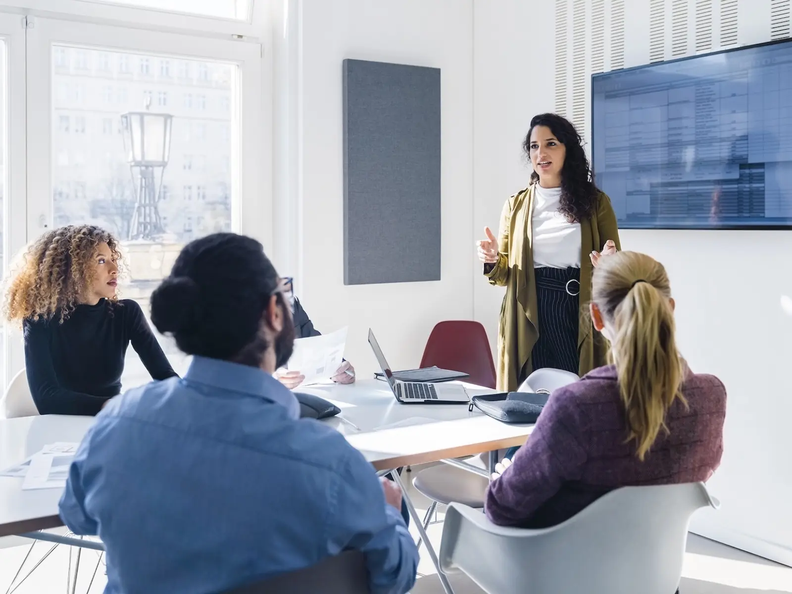 A diverse group in a bright conference room listens attentively as a woman presents information, with a laptop and documents on the table.