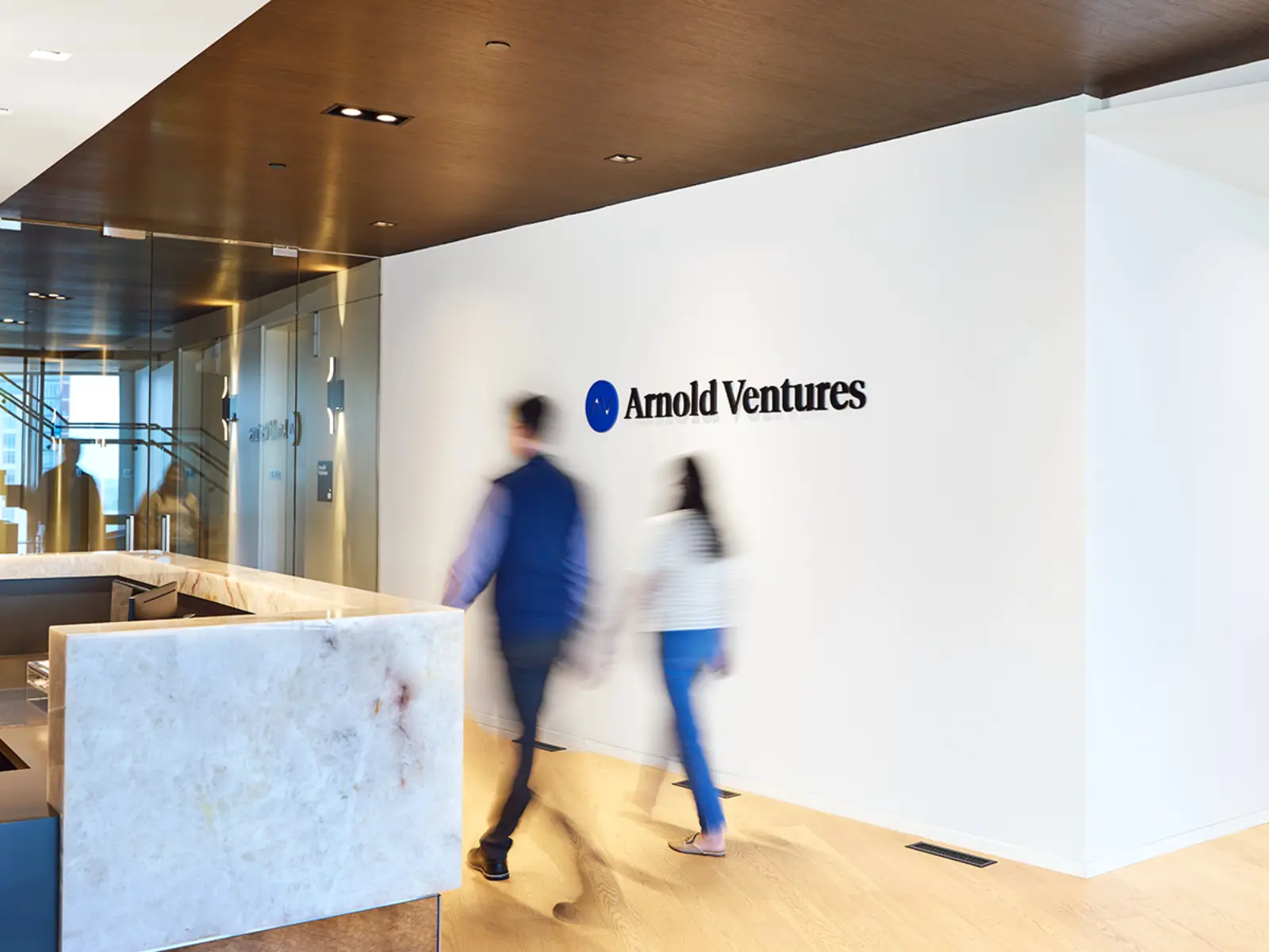 Two people walk through a modern office lobby, showcasing a sleek reception area with a prominent “Arnold Ventures” logo on the wall.