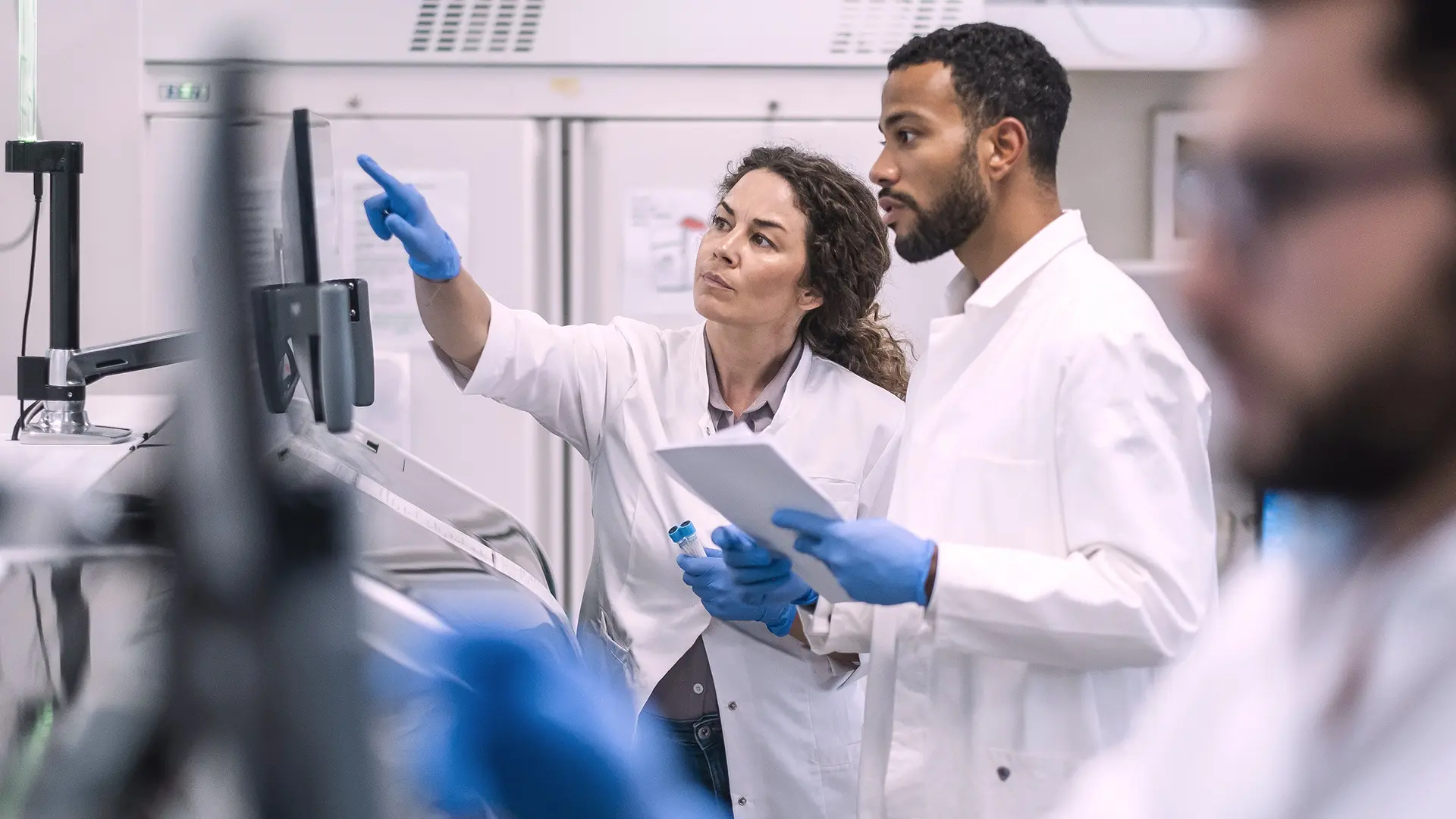 Two scientists in white lab coats examine data on a computer screen, one pointing and the other holding a notepad, deep in discussion.