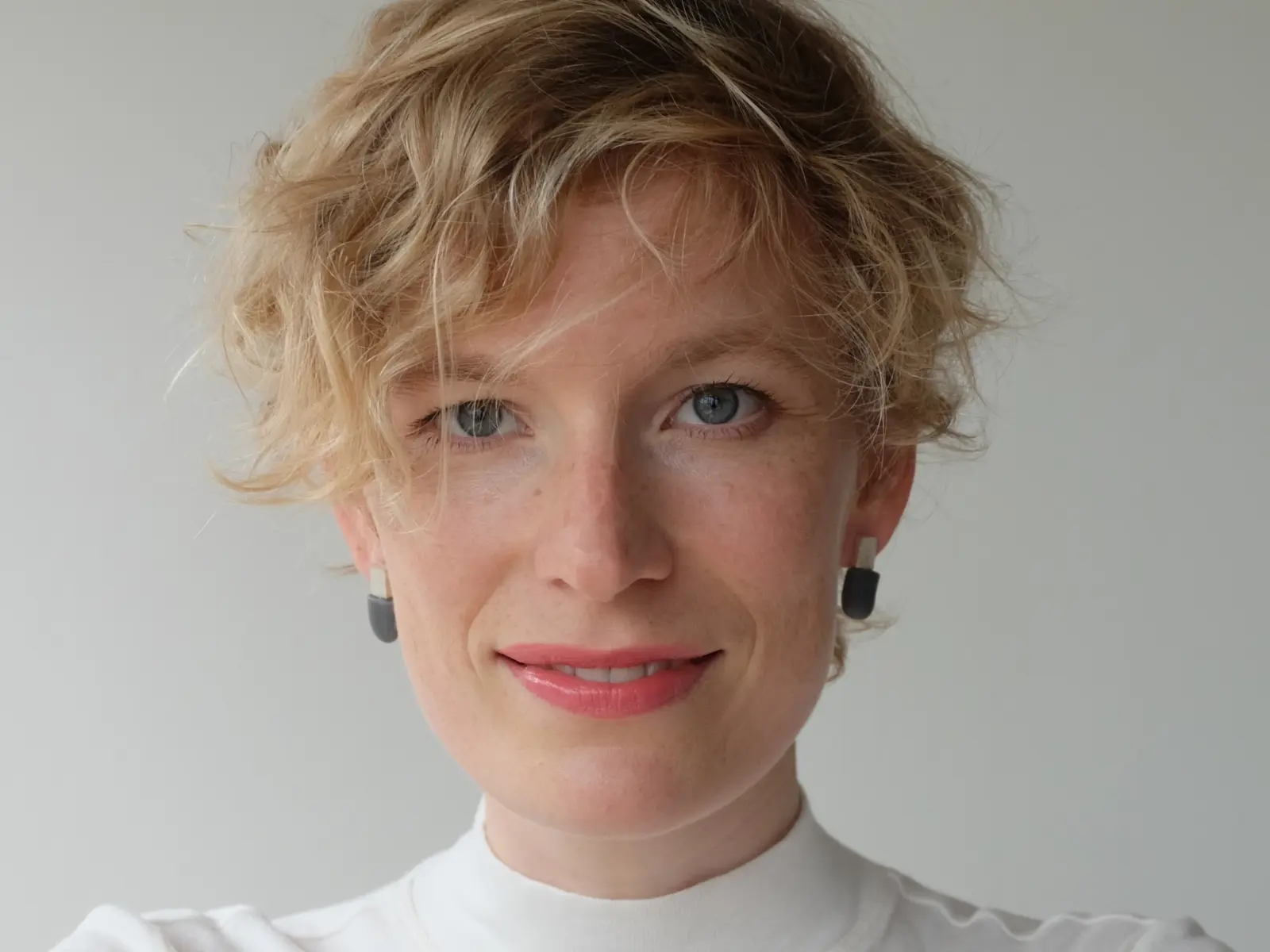 A smiling person with short, curly blonde hair wears a white turtleneck and black earrings, against a light background.