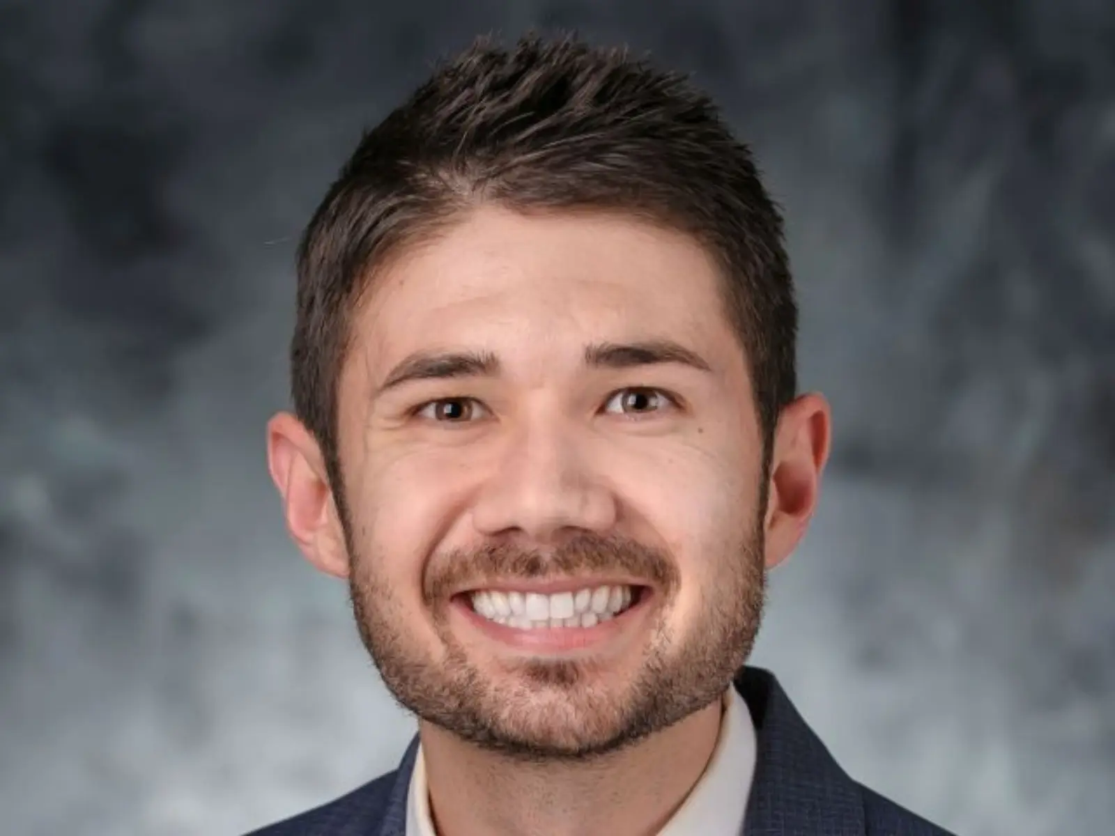 Smiling man with dark hair, wearing a blue suit and tie, against a soft gray backdrop.