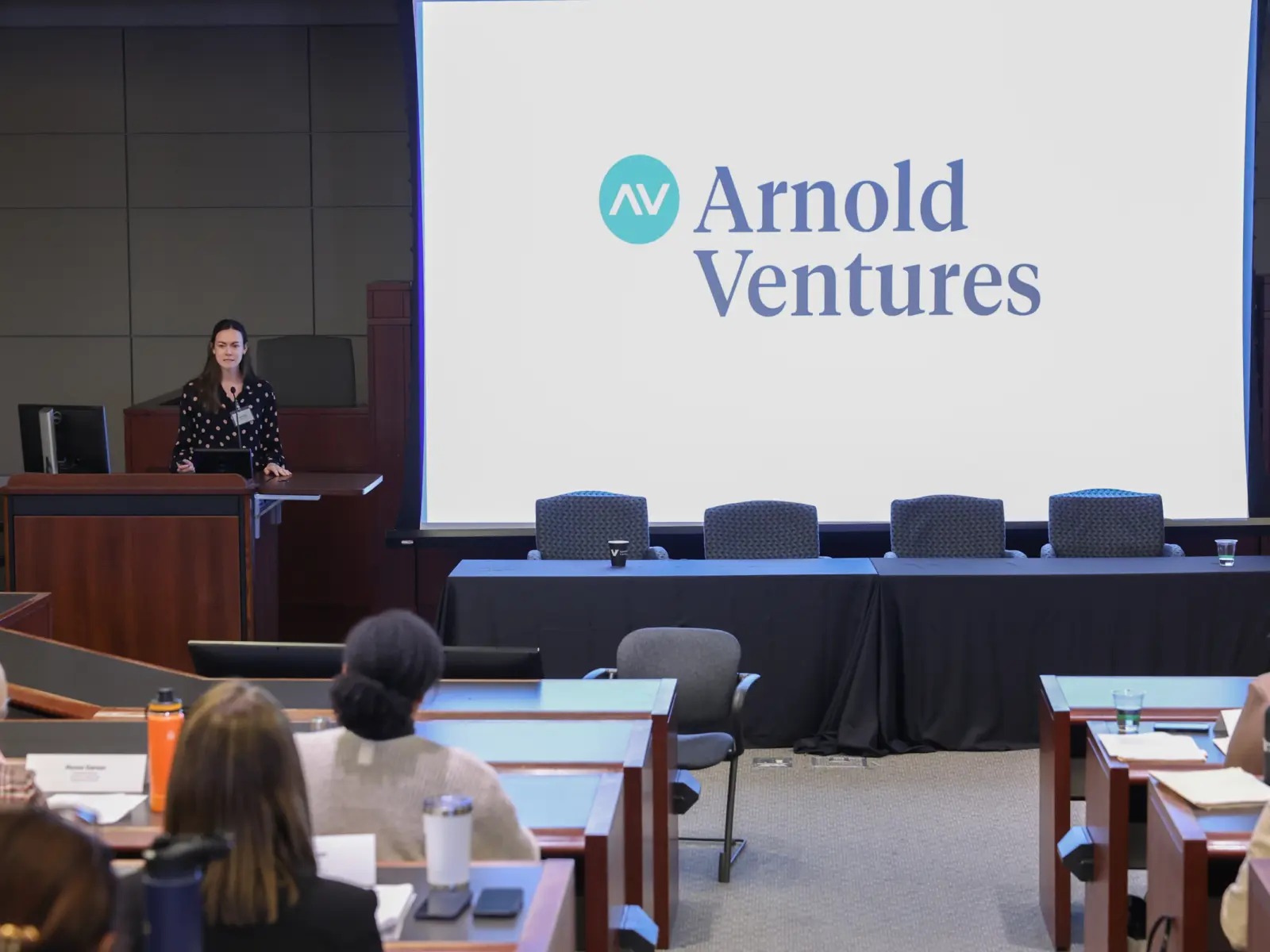 A woman stands at a podium addressing an audience, with a large screen displaying the Arnold Ventures logo behind her.