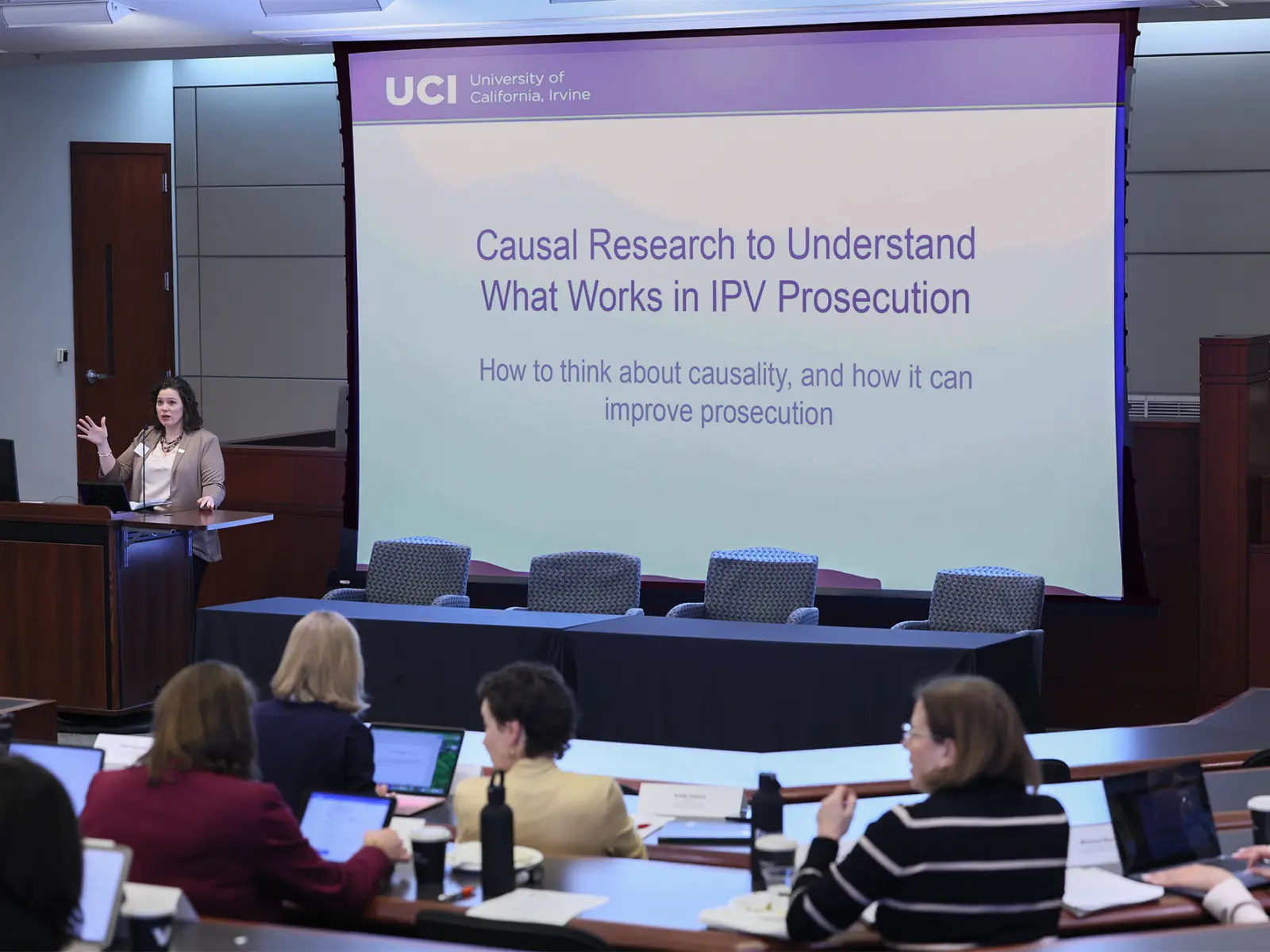 A presenter stands at a podium explaining the role of causal research in improving IPV prosecution, with attendees engaged.