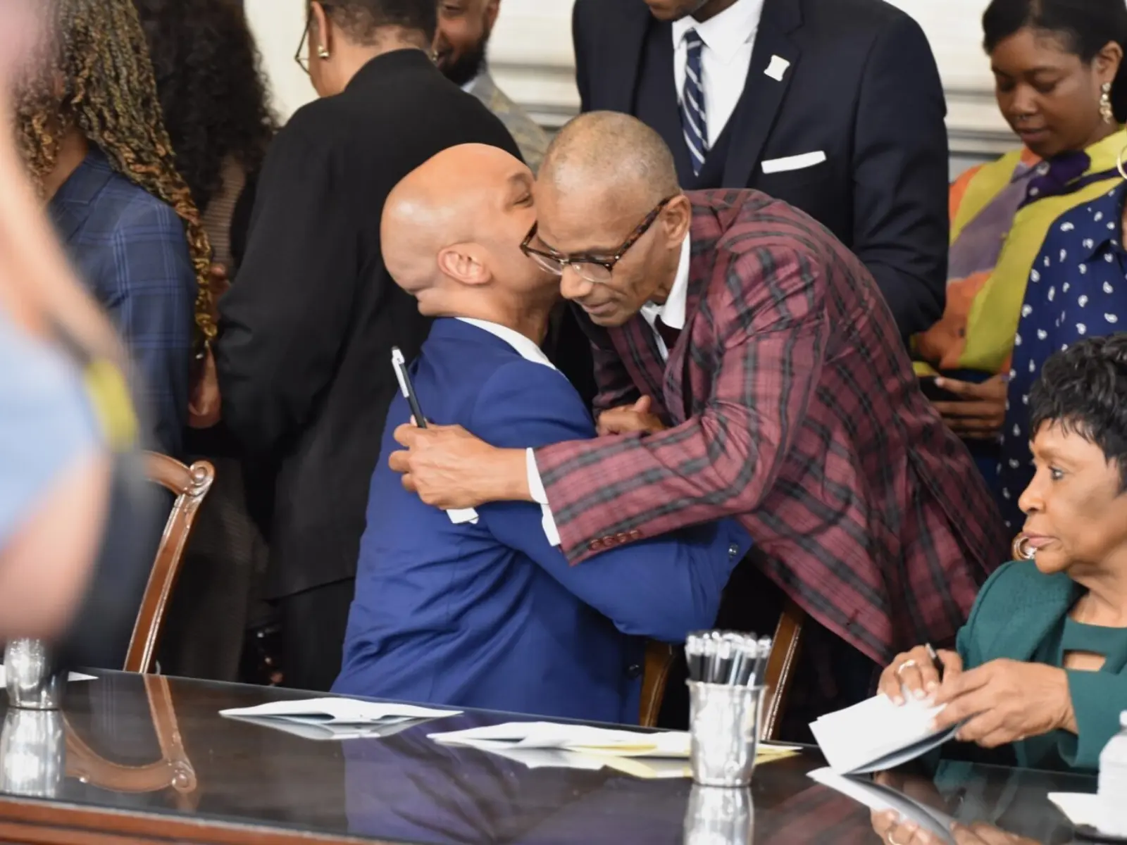Two men embrace warmly at a formal event, surrounded by attendees. One man is dressed in a blue suit, the other in a plaid jacket.