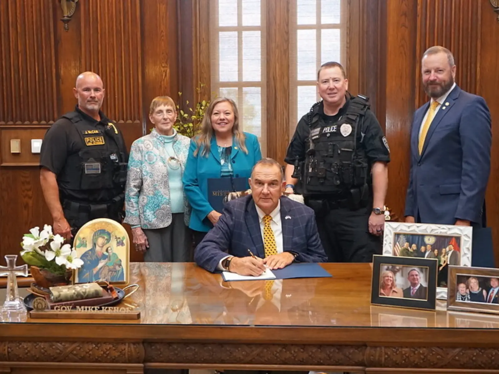A group of six individuals gathers around a desk in an office, with one person signing documents while others stand nearby, smiling.