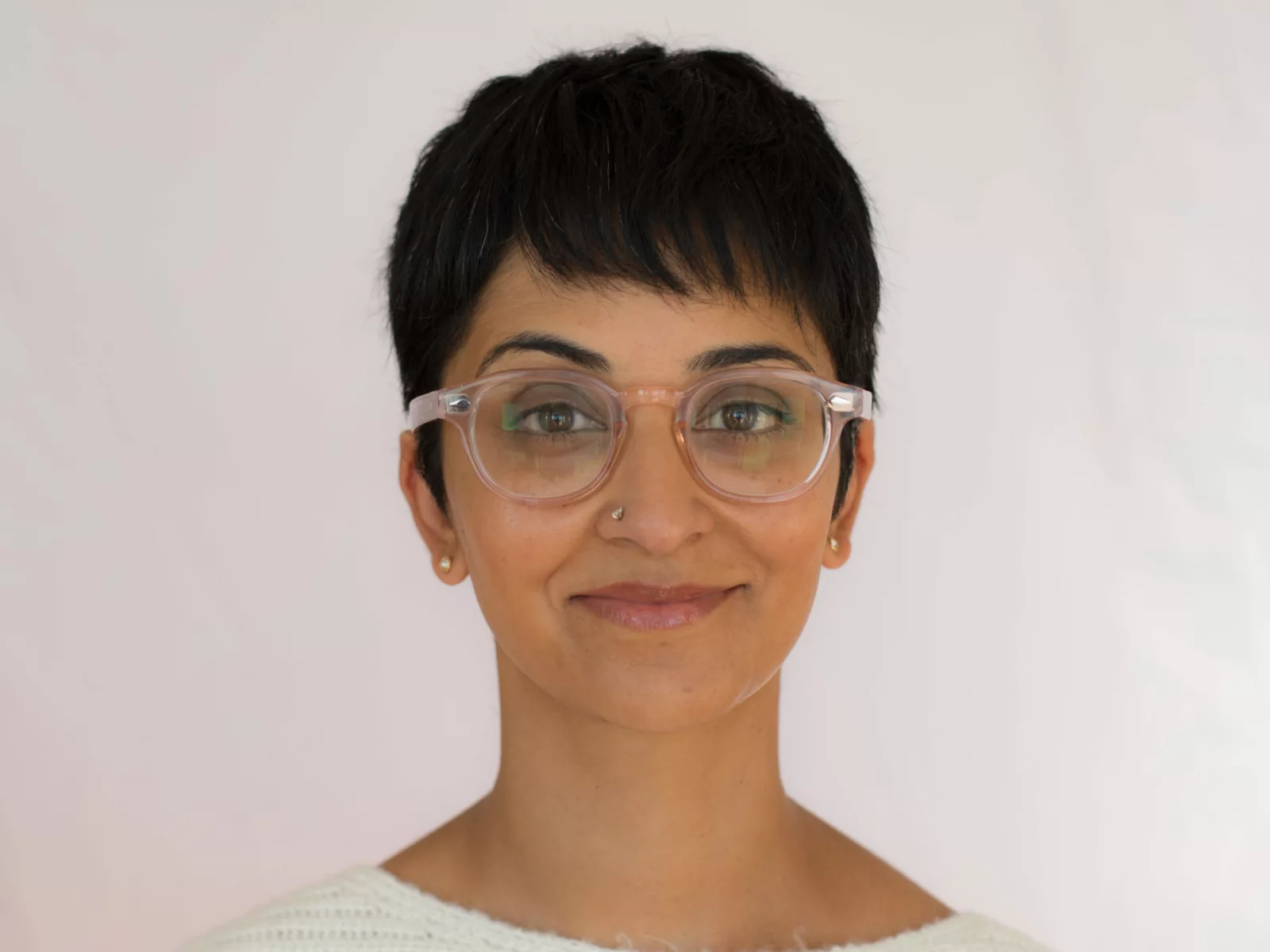 A smiling person with short, dark hair and glasses, wearing a cozy white sweater, stands against a soft, light background.