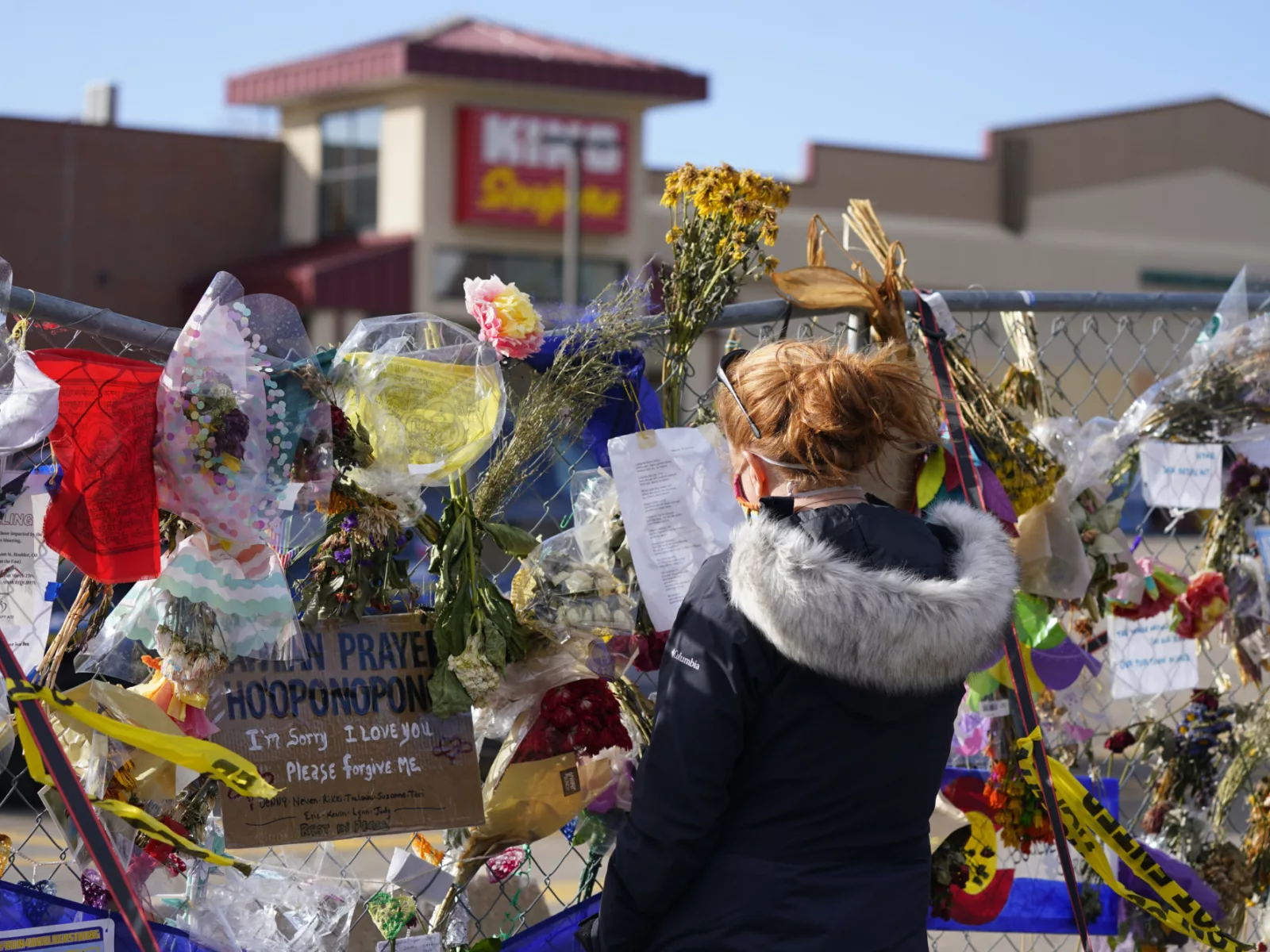 A woman stands before a colorful memorial on a fence, adorned with flowers, notes, and heartfelt messages of love and remembrance.