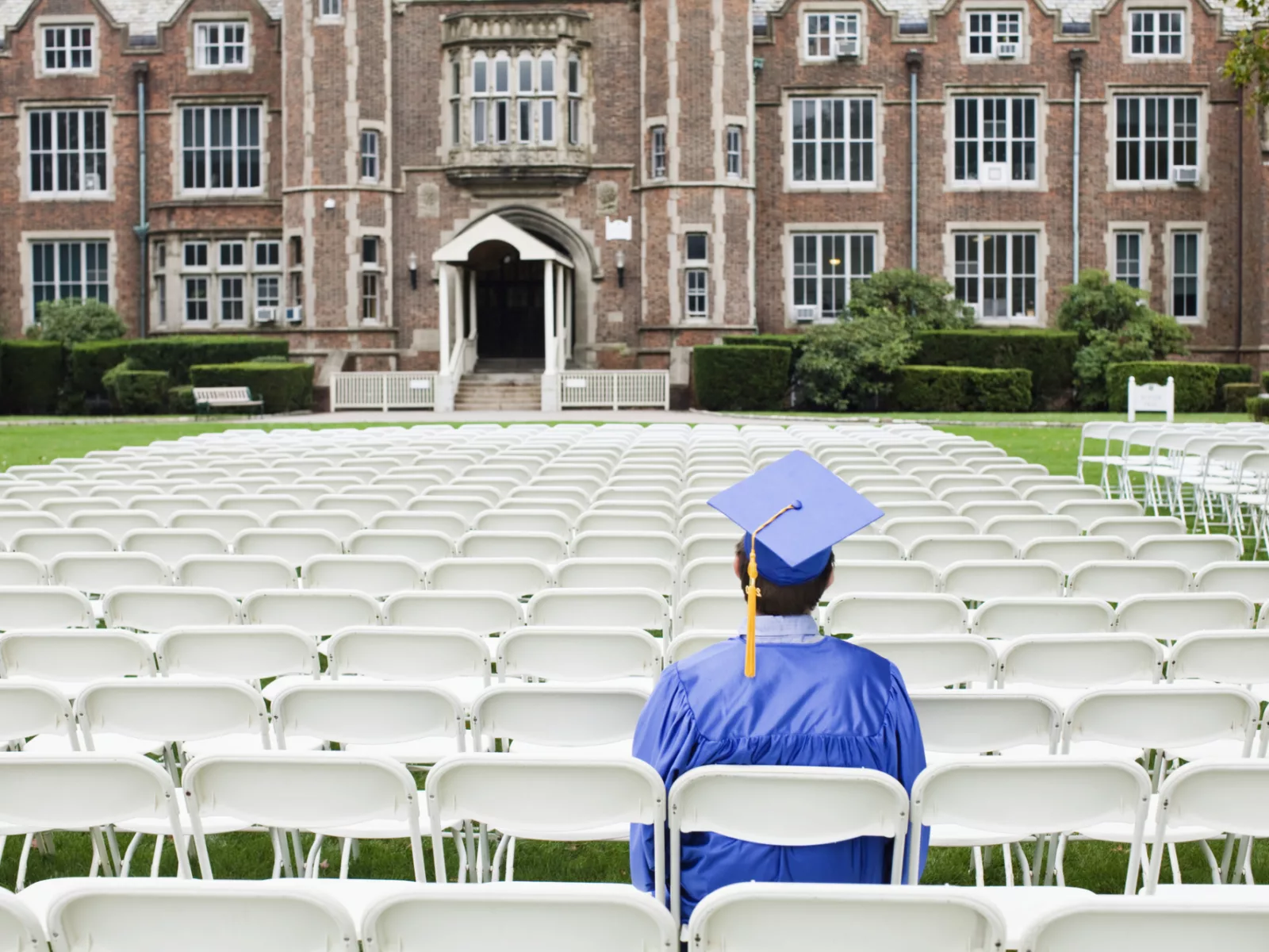 A graduate in a blue cap and gown sits among empty white chairs, facing a historic brick building, ready for the commencement ceremony.
