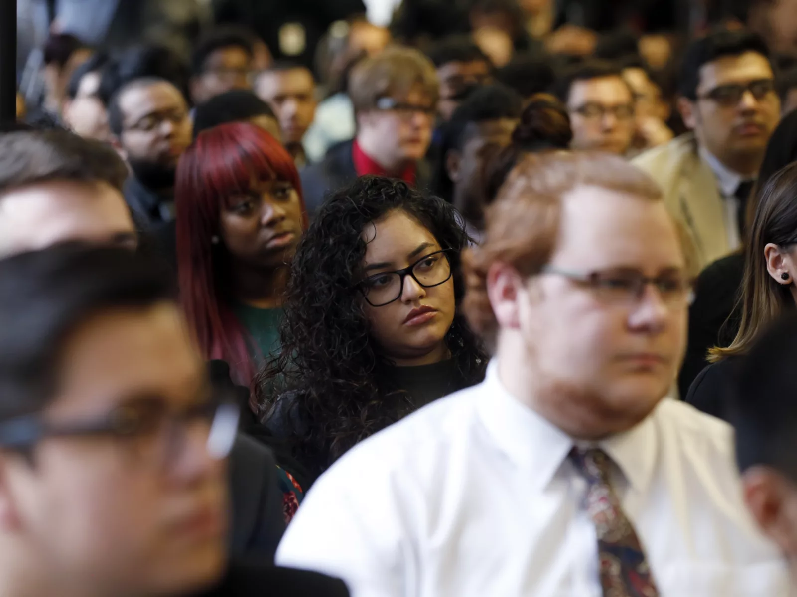 A diverse group of people sits attentively, with focused expressions. The foreground features individuals wearing glasses and formal attire.