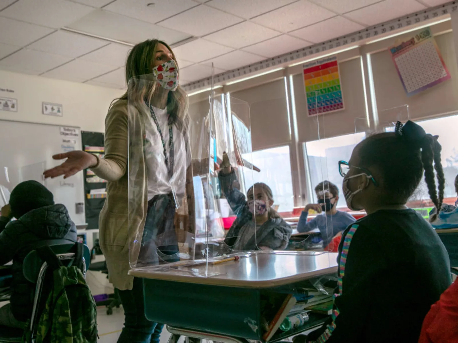 Teacher wearing mask stands at front of classroom with students sitting at desks.