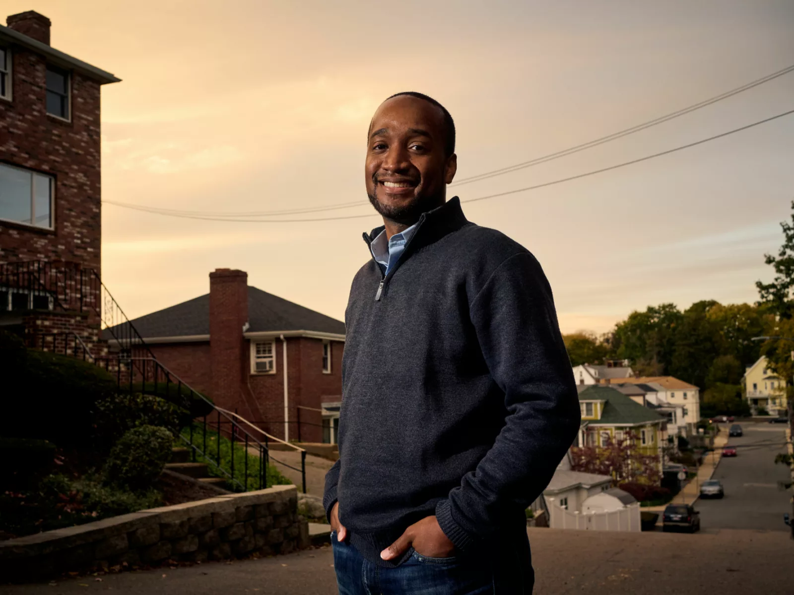 A smiling man stands confidently on a sloped street, dressed in a dark zip-up sweater, with houses and trees in the background.