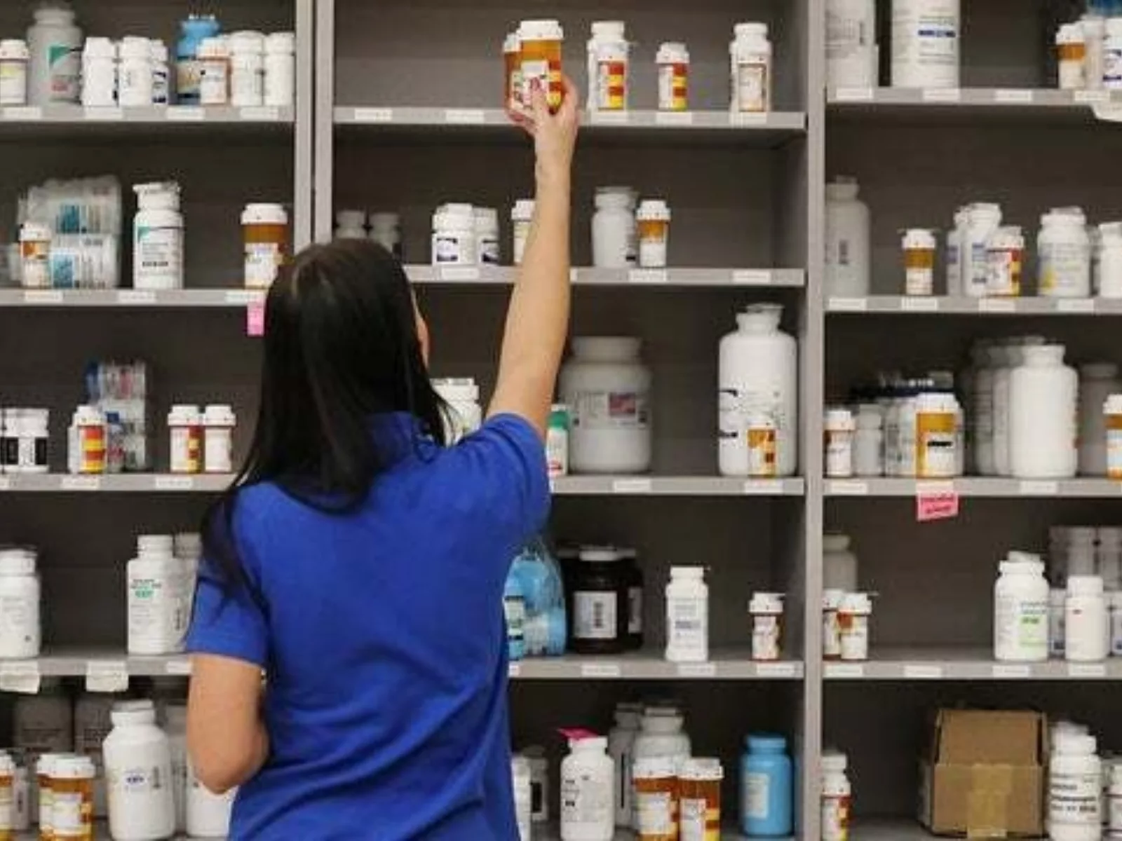 A pharmacy employee in a blue shirt reaches for a prescription bottle on a shelf filled with various labeled medicine containers.