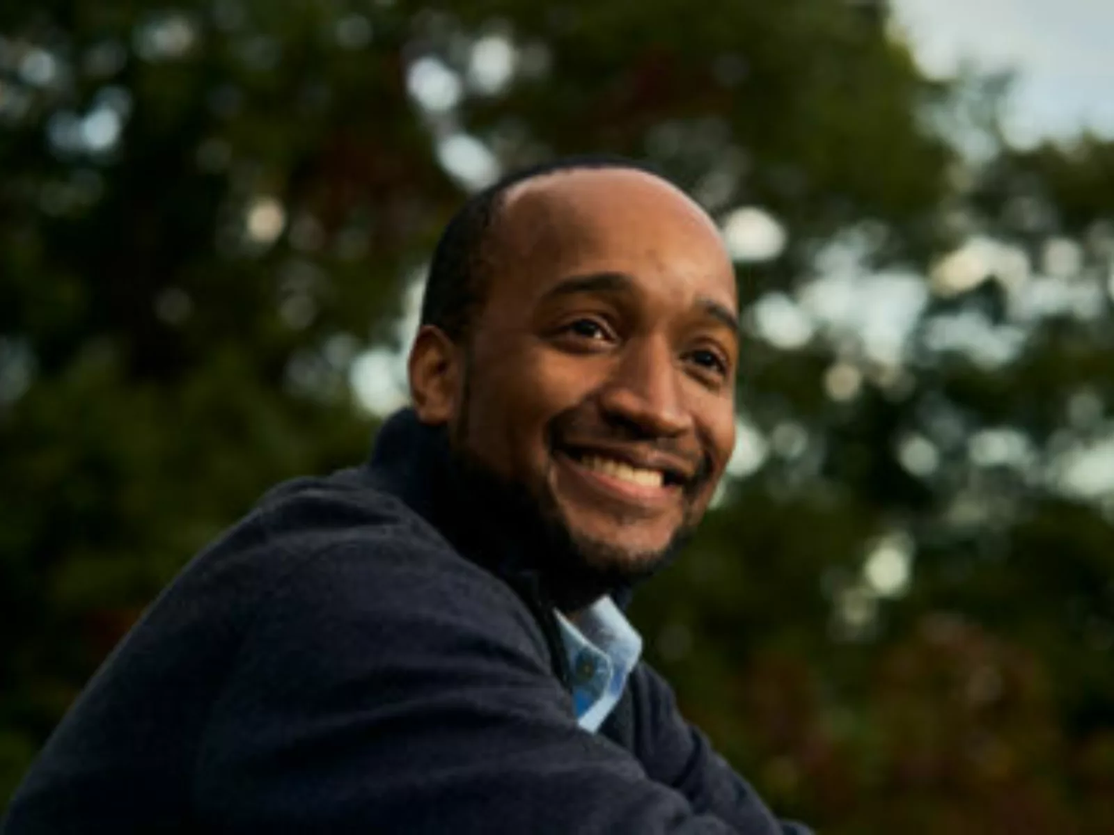 A man with a warm smile sits outdoors, wearing a dark sweater, with trees blurred in the background, capturing a serene moment.